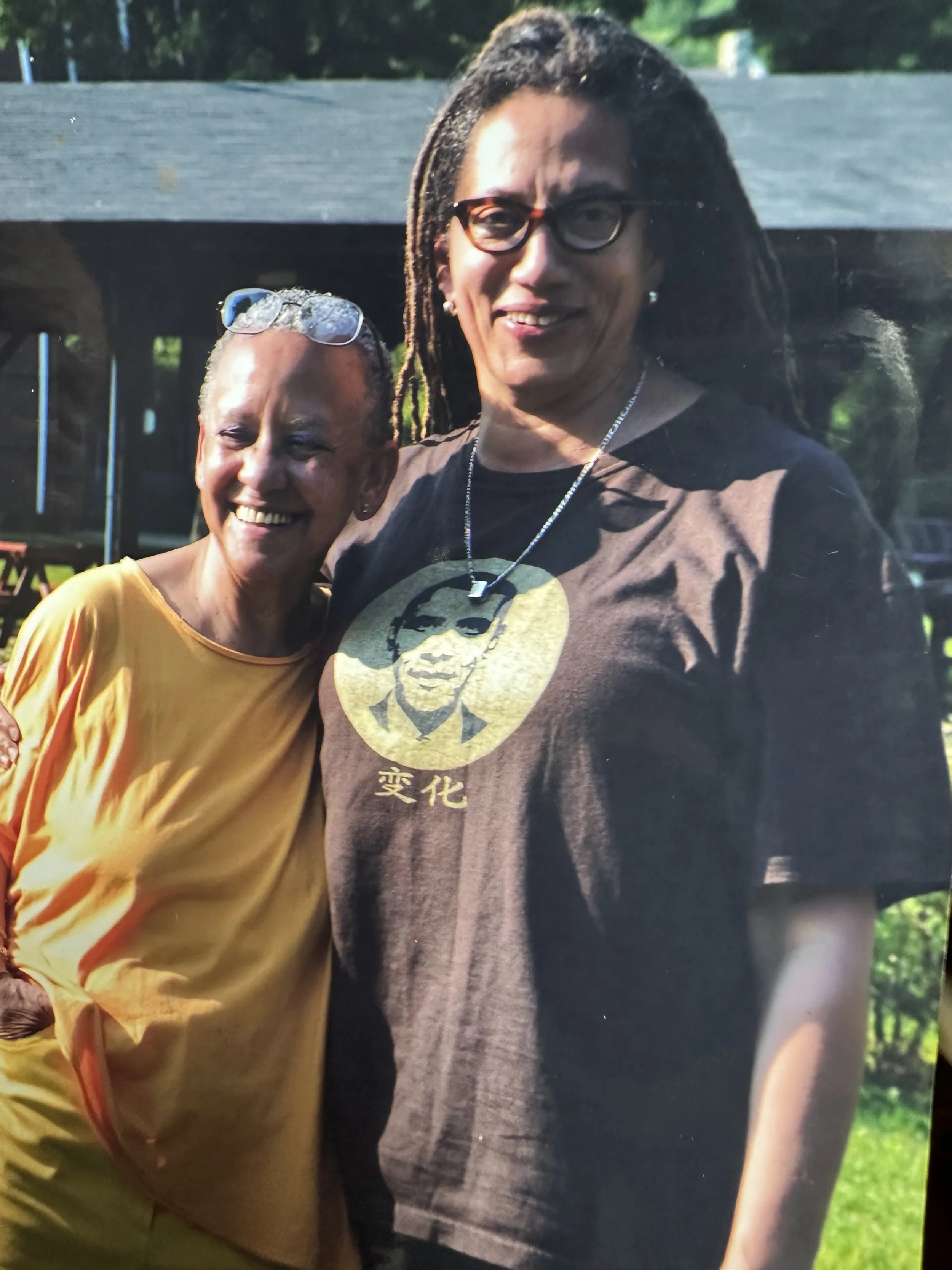 Nikky Finney and Nikki Giovanni, standing outdoors, smiling and embracing each other, with trees and a fence in the background.