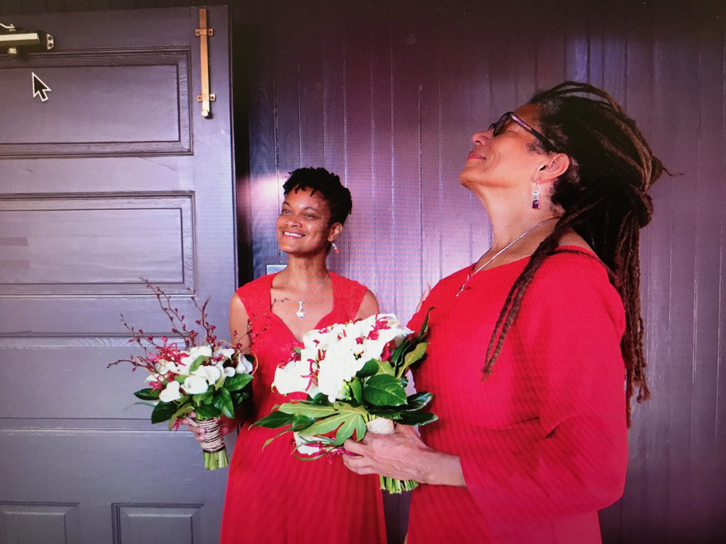 A woman with long dreadlocks wearing glasses and a red dress holding a large bouquet of white and pink flowers with green leaves, smiling and standing in front of a purple wall with vertical striped pattern.