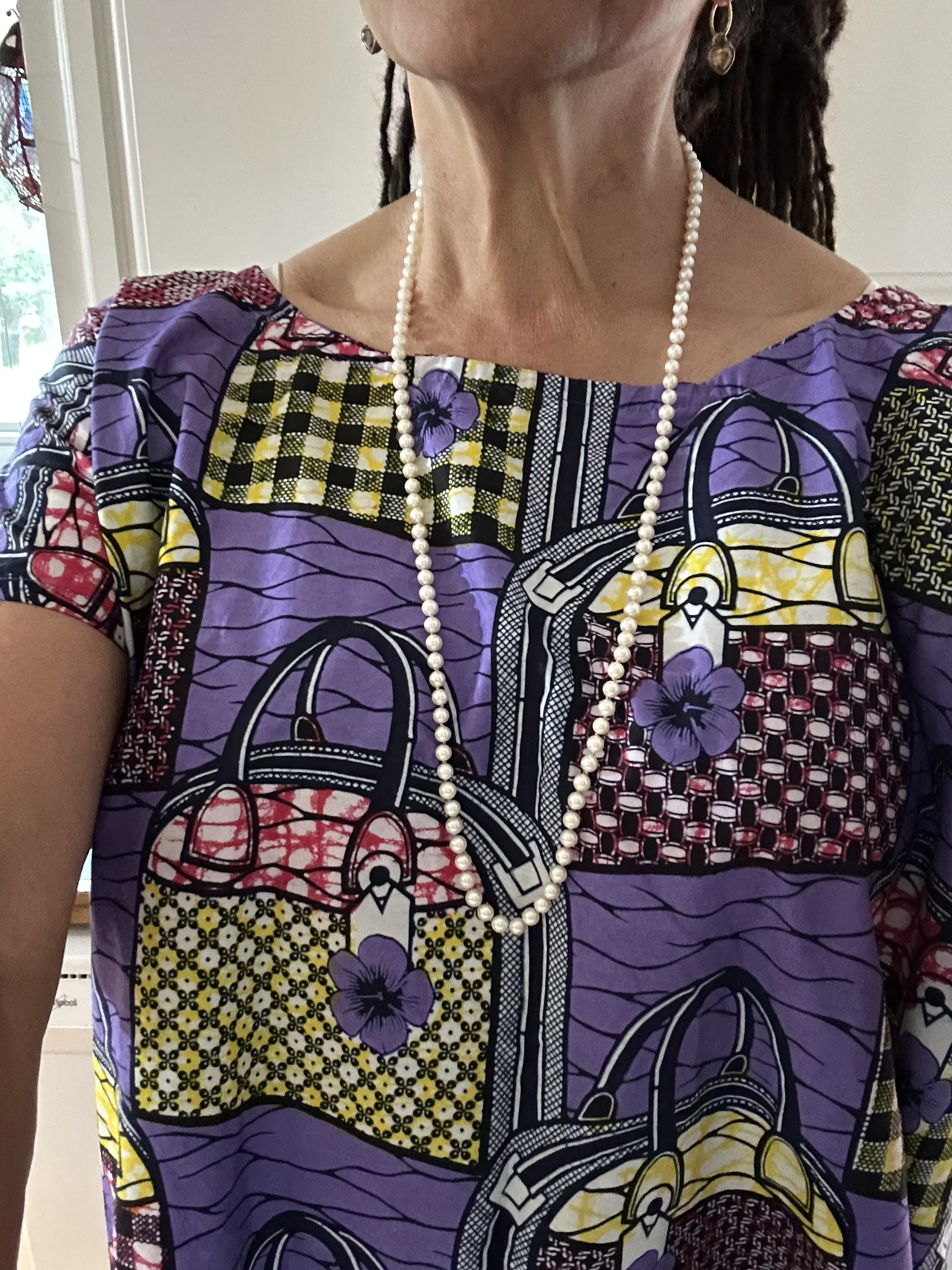Close-up of Nikky Finney wearing a colorful patterned dress with purple, yellow, red, and black designs, a pearl necklace, and earrings. Part of her face and shoulder are visible, and she has dreadlocks. 