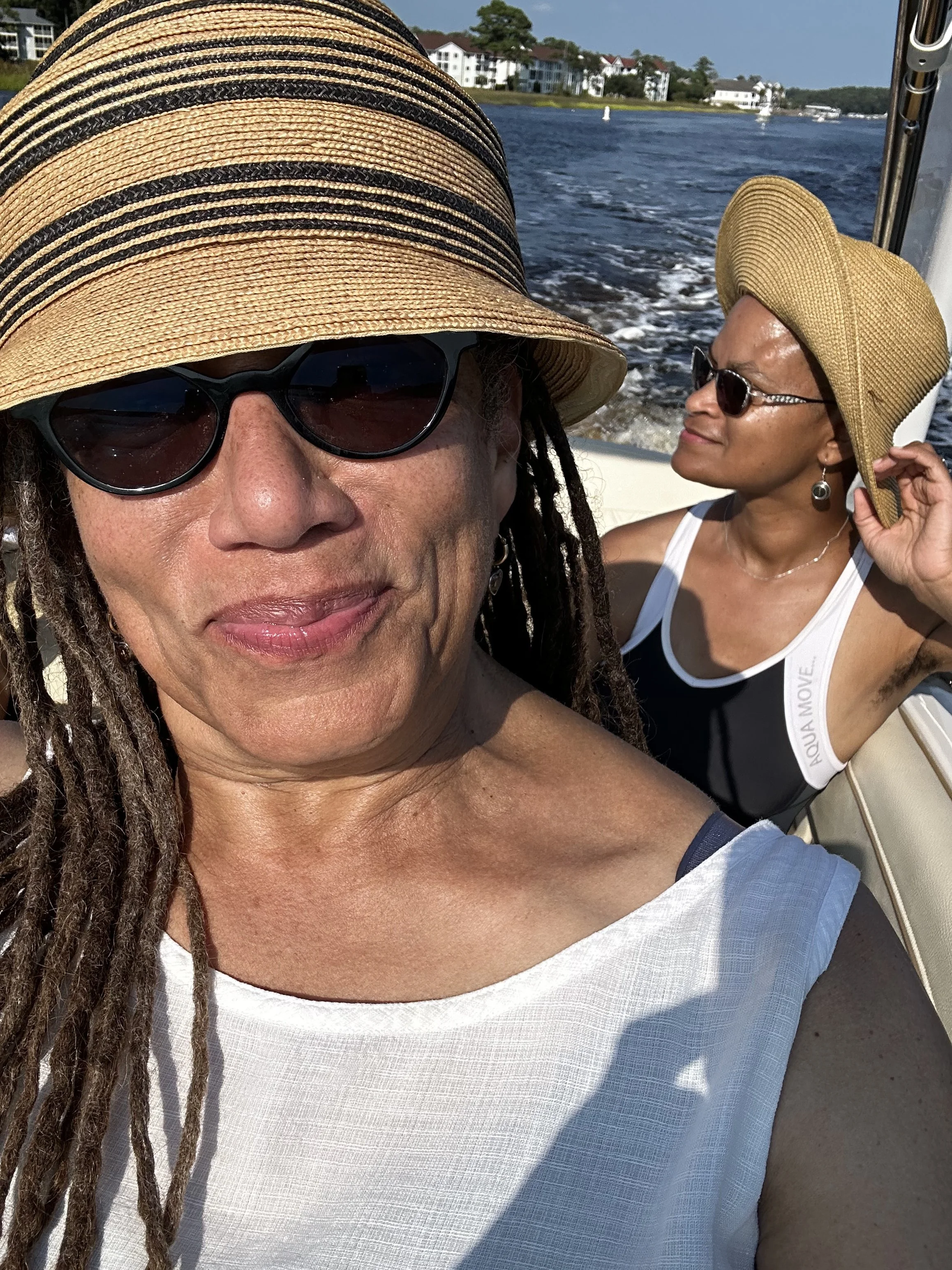 Nikky Finney & Tonia Poteat wearing sun hats and sunglasses enjoying a boat ride on a sunny day, with houses and water in the background.