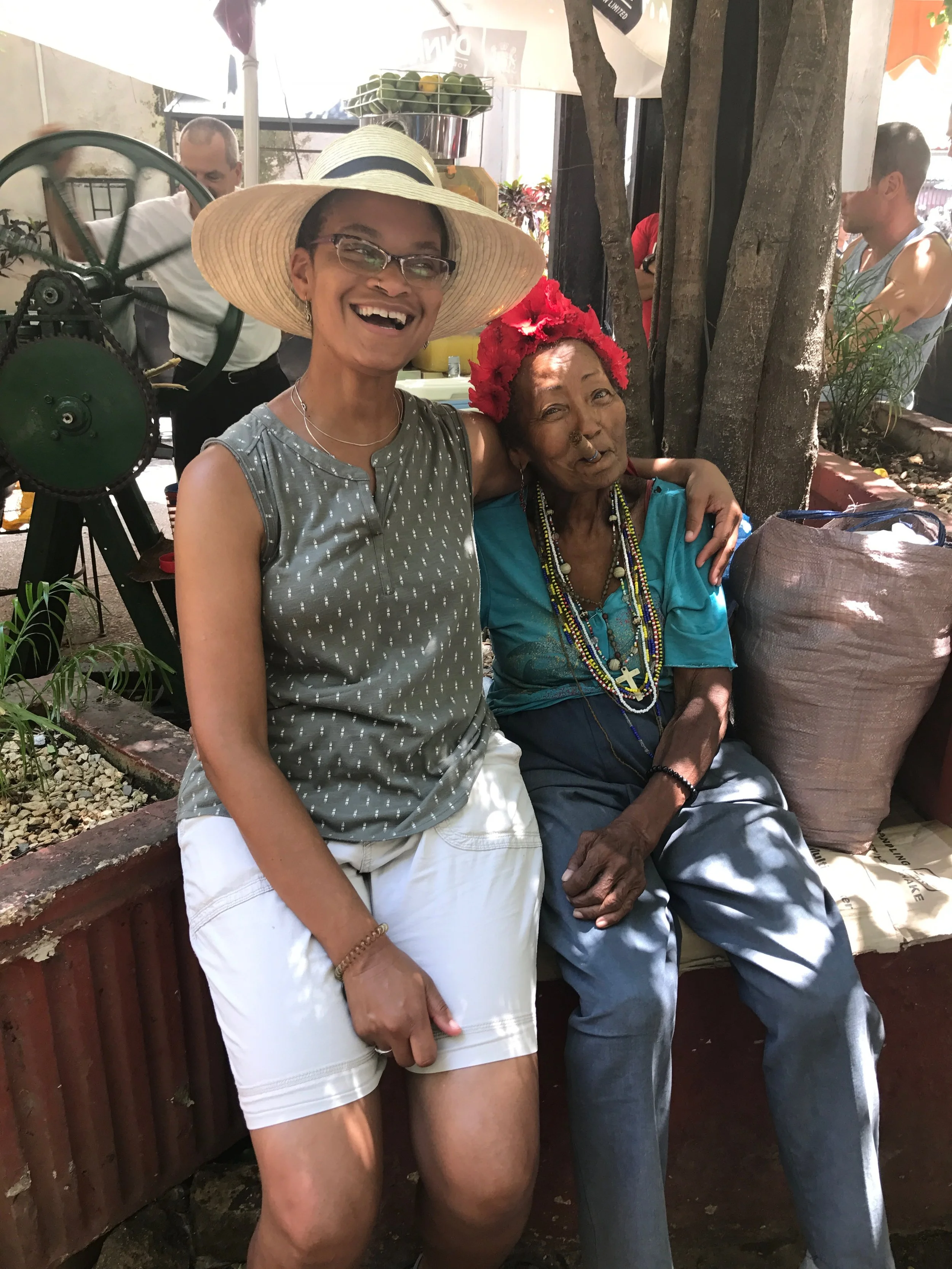 Two women sitting outdoors, smiling. The woman on the left, wearing glasses, a wide-brimmed hat, and a sleeveless top, is standing next to another woman who has a red floral headpiece, some beaded necklaces, and is wearing a blue shirt.
