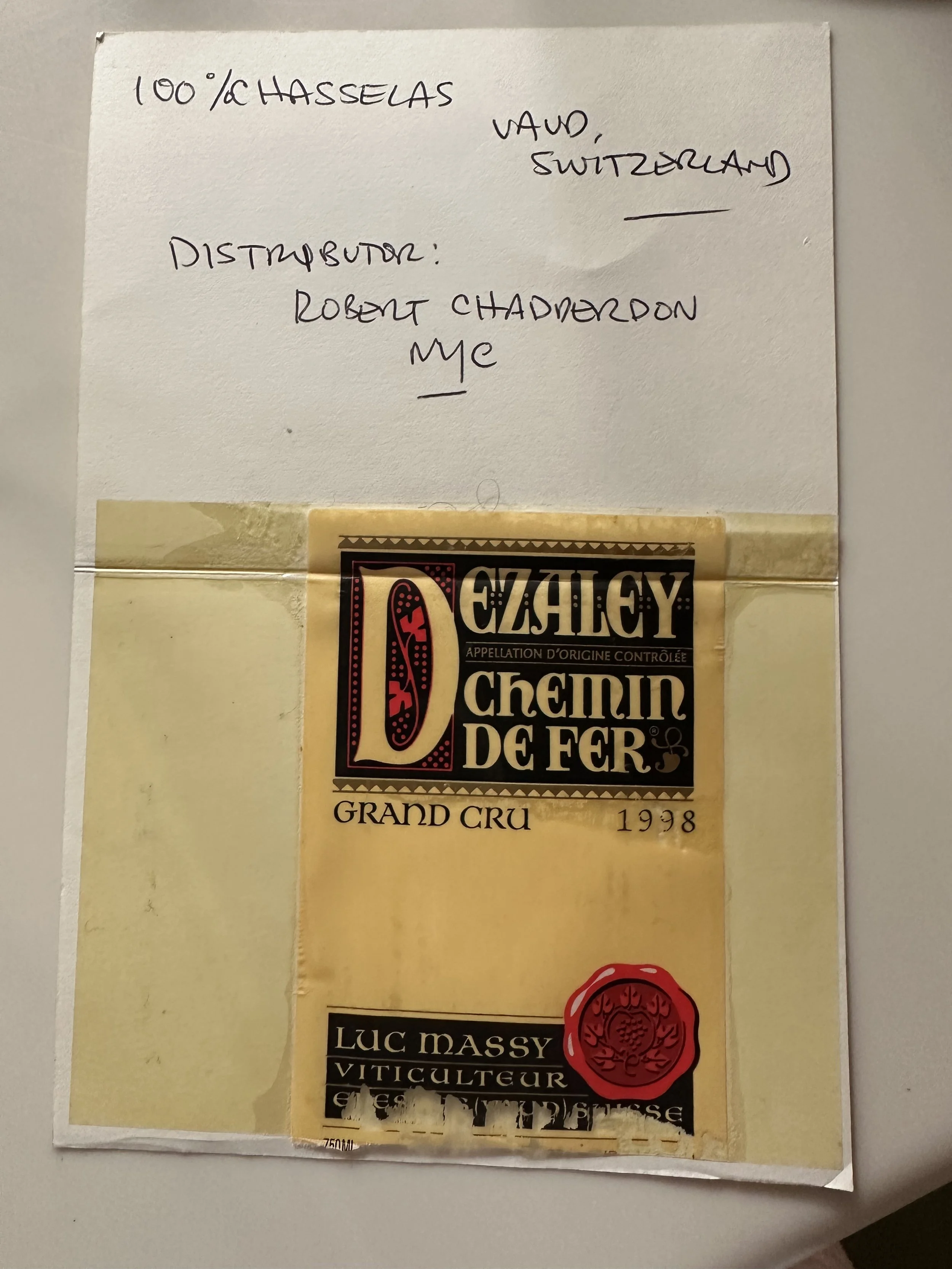 Handwritten note on white paper indicates the wine is 100% Haselras from Vaud, Switzerland. It is labeled as a grand cru from 1998 and is called Chemin de Fer. Part of a wine label is visible beneath the note.