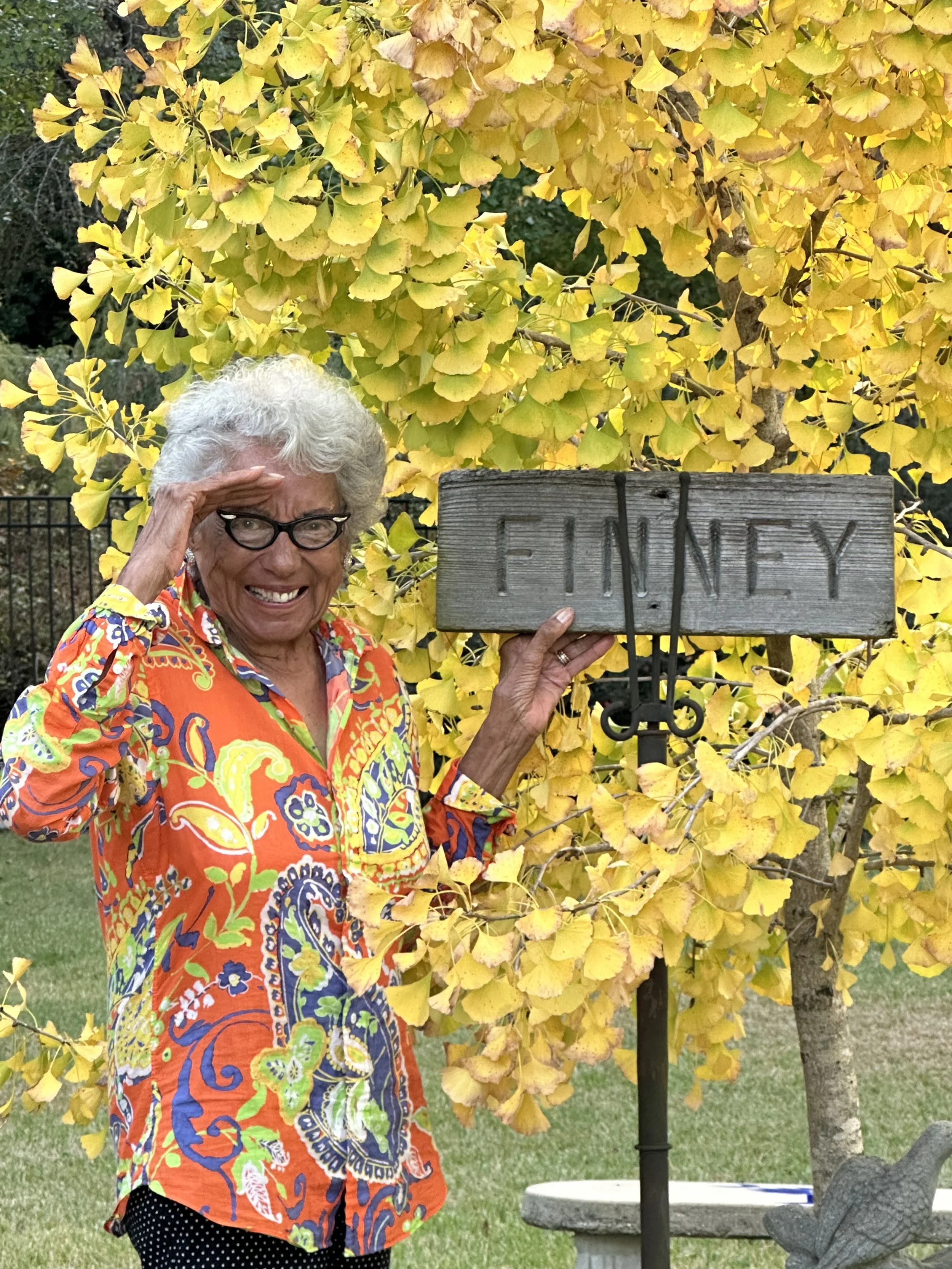 An elderly woman with curly gray hair and glasses, wearing a colorful, patterned shirt, is standing outdoors next to a yellow ginkgo tree and holding a wooden sign with the word 'FINEY' written on it. She is smiling and saluting with her right hand.