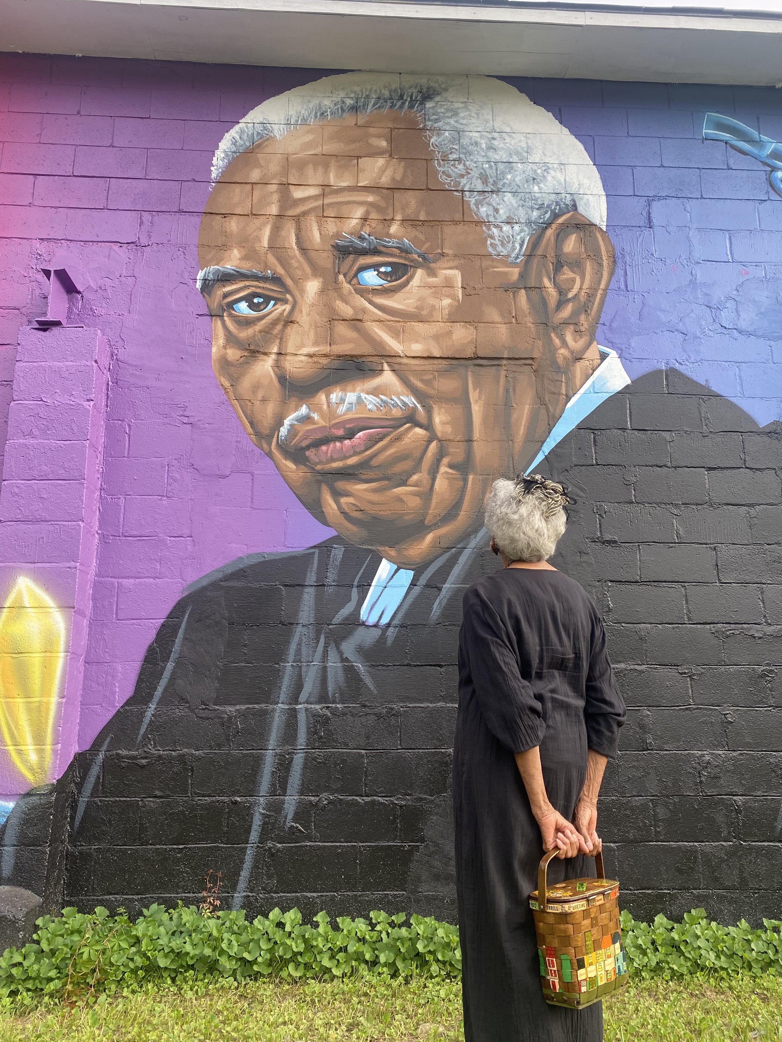 A mural of Earnest A Finney, Jr. painted on a brick wall, with an elderly woman with white hair looking at it, holding a shopping basket and standing in front of green plants and purple wall backgrounds.