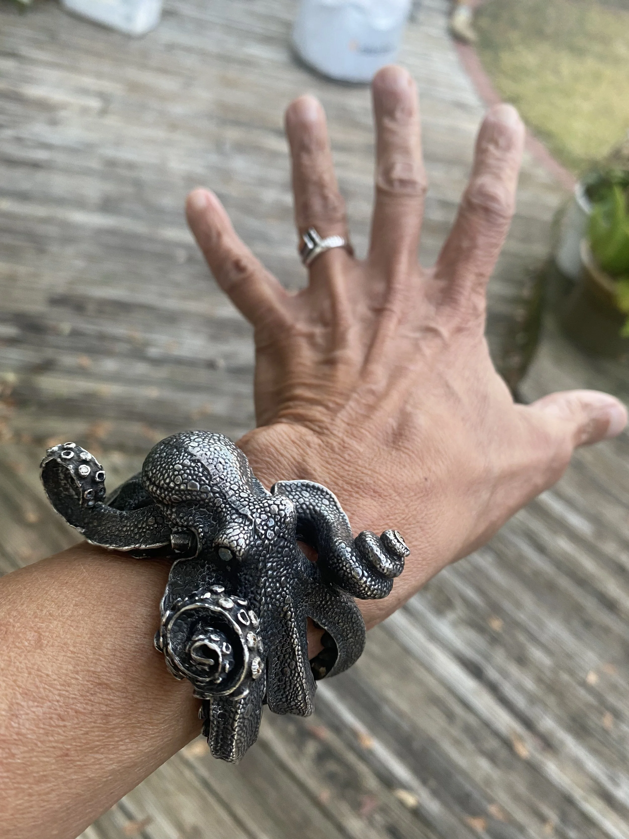 A hand with a silver ring on the middle finger shows a black and silver octopus-shaped bracelet, with a blurred wooden deck background and some potted plants.