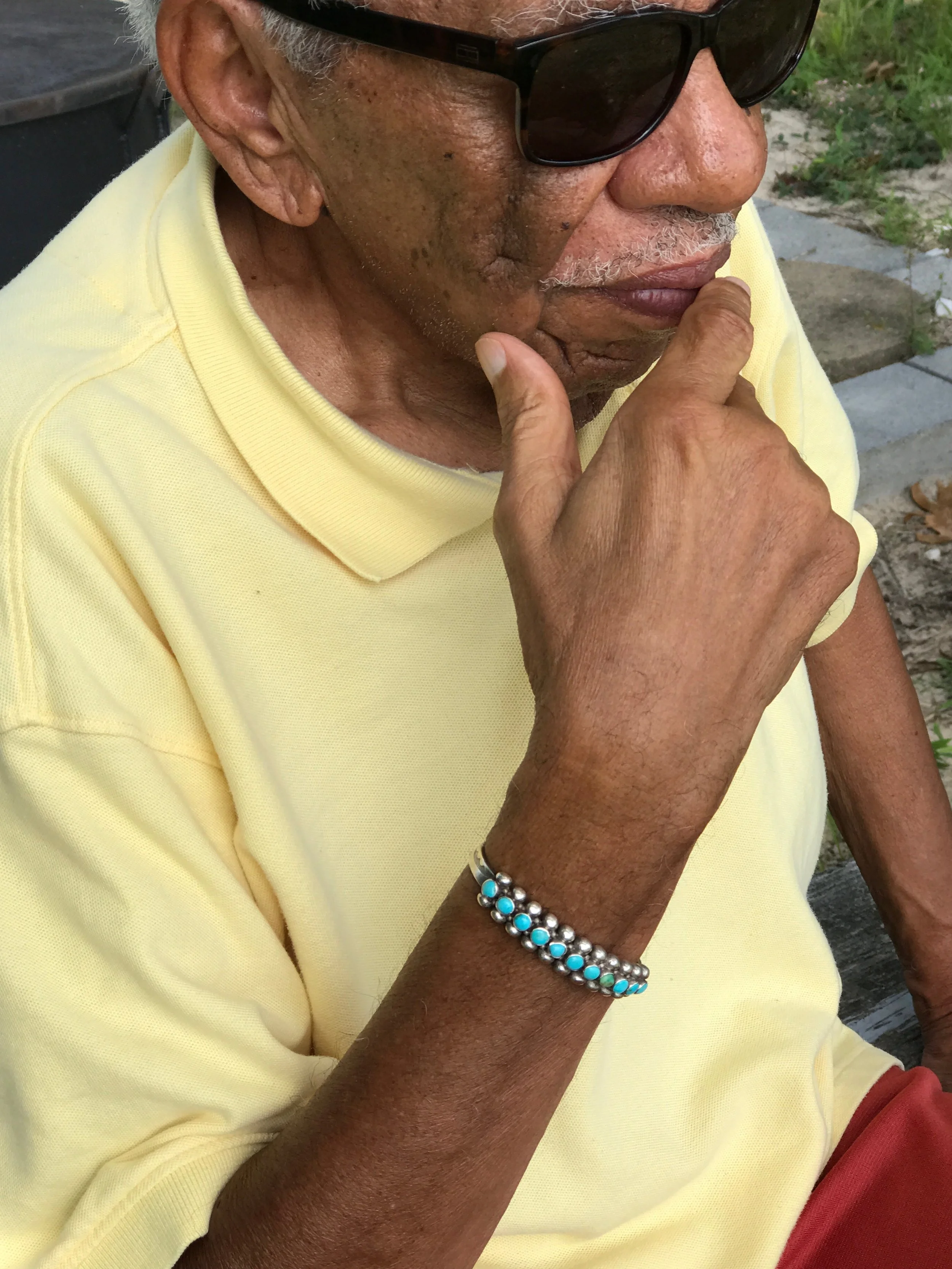 Close-up of an elderly man with dark skin wearing sunglasses, a yellow shirt, and a turquoise beaded bracelet, sitting outdoors with grass and stone pavement in the background, holding his chin in a thinking pose.