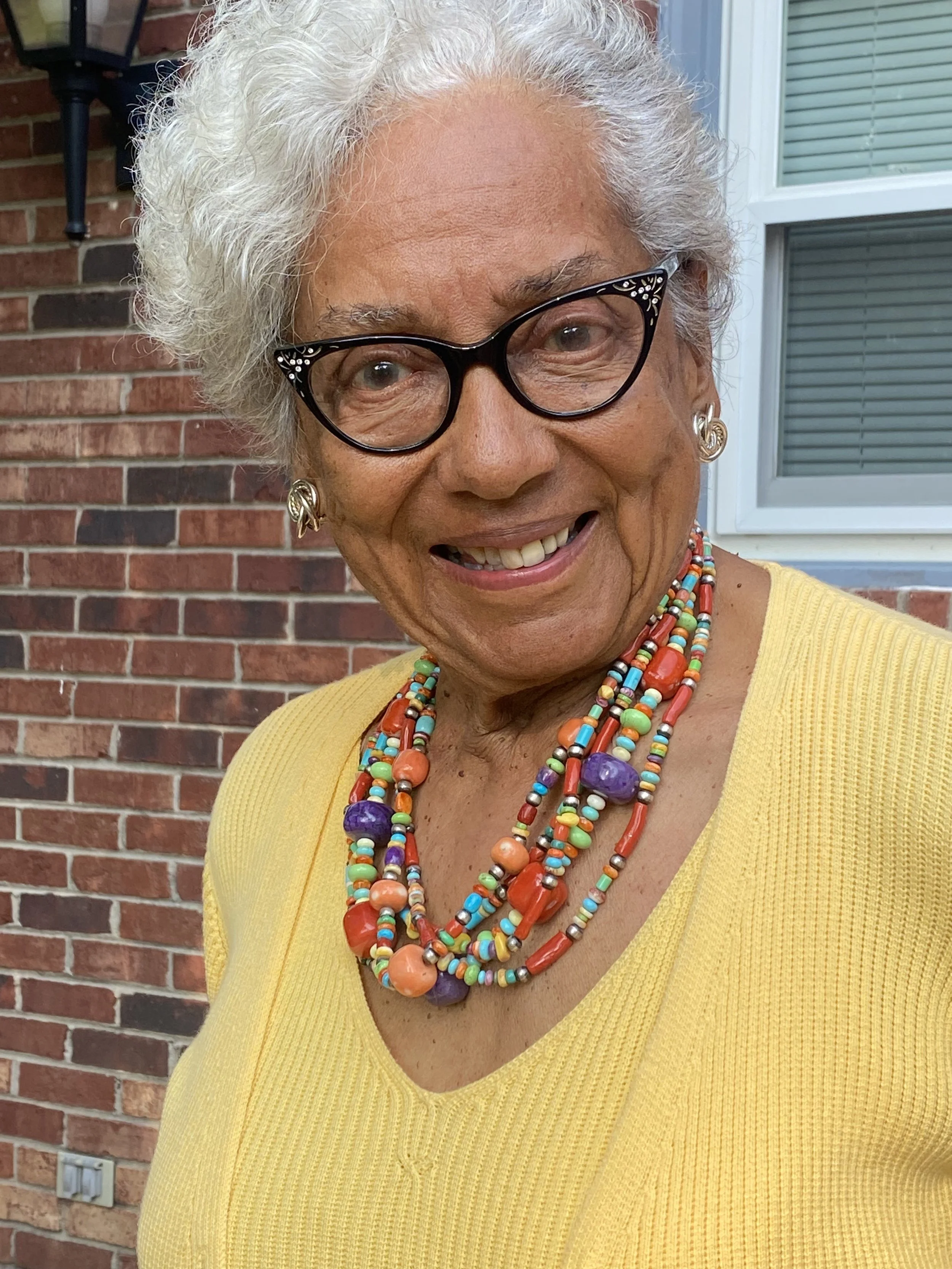 Close-up of an older woman with curly white hair wearing black glasses, gold earrings, colorful beaded necklaces, a yellow sweater, and standing in front of a brick wall and window.