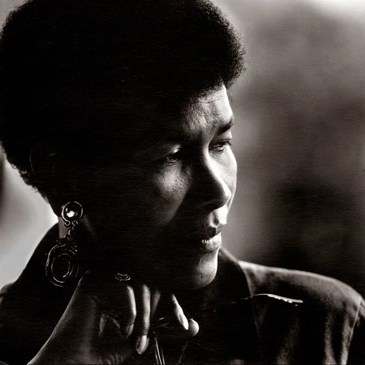 A black and white portrait of a woman with short, natural hair, wearing earrings, with a thoughtful expression, resting her chin on her hand.
