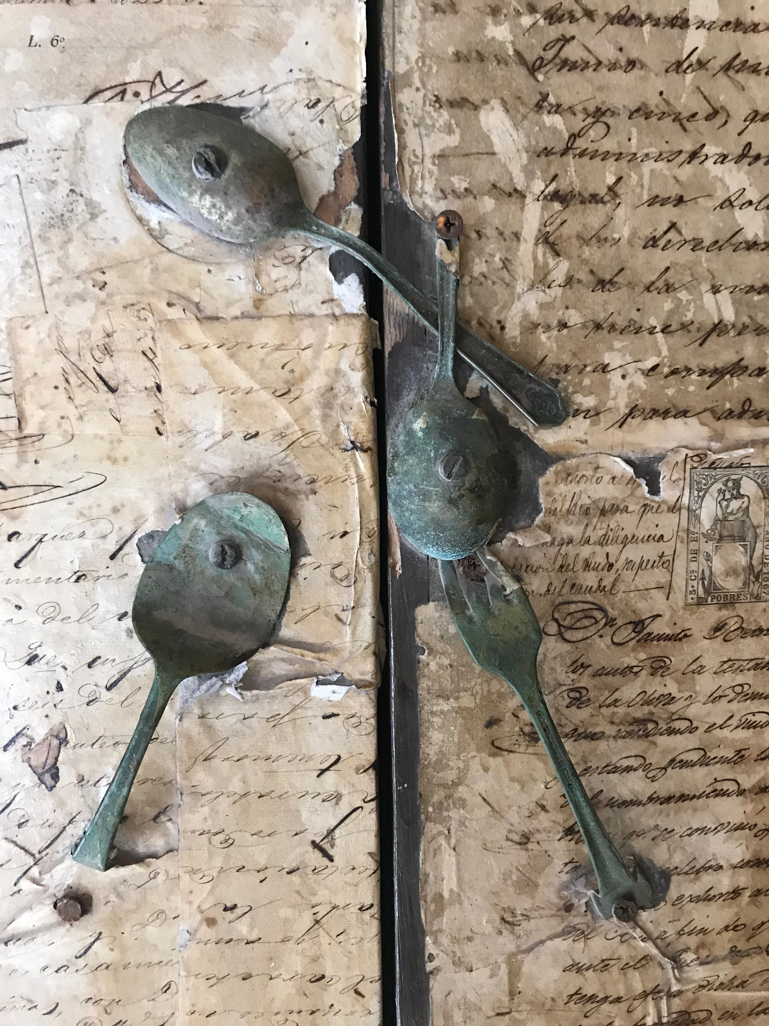 A close-up image of two tarnished, greenish-brown metal spoons on what appears to be an aged, torn handwritten letter or manuscript with elegant cursive script. The background paper is yellowed and damaged.