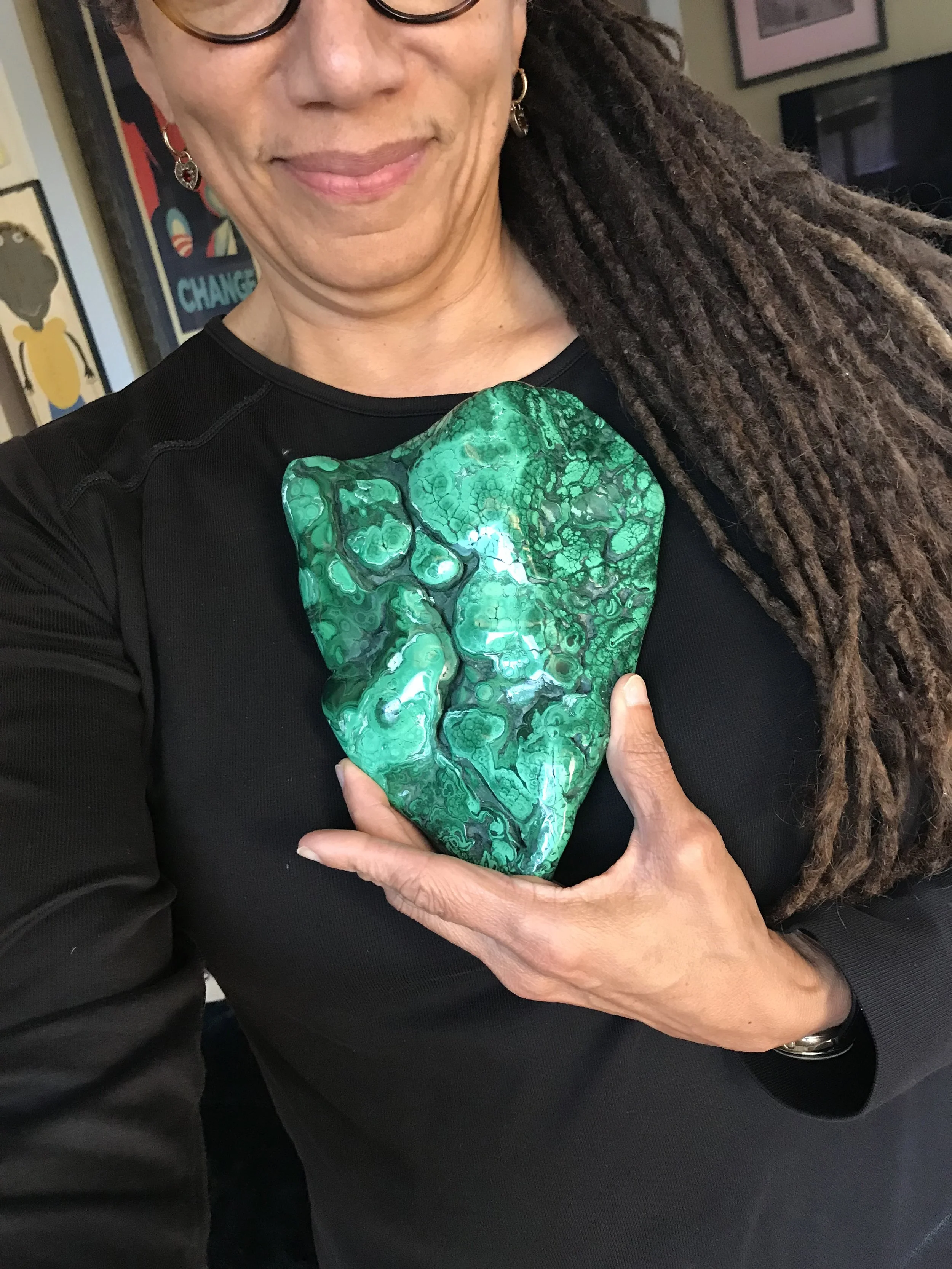Nikky Finney holding a large, green, heart-shaped gemstone or mineral with a bubbly, textured appearance, wearing a black top and glasses, with a background of framed artwork and a television.