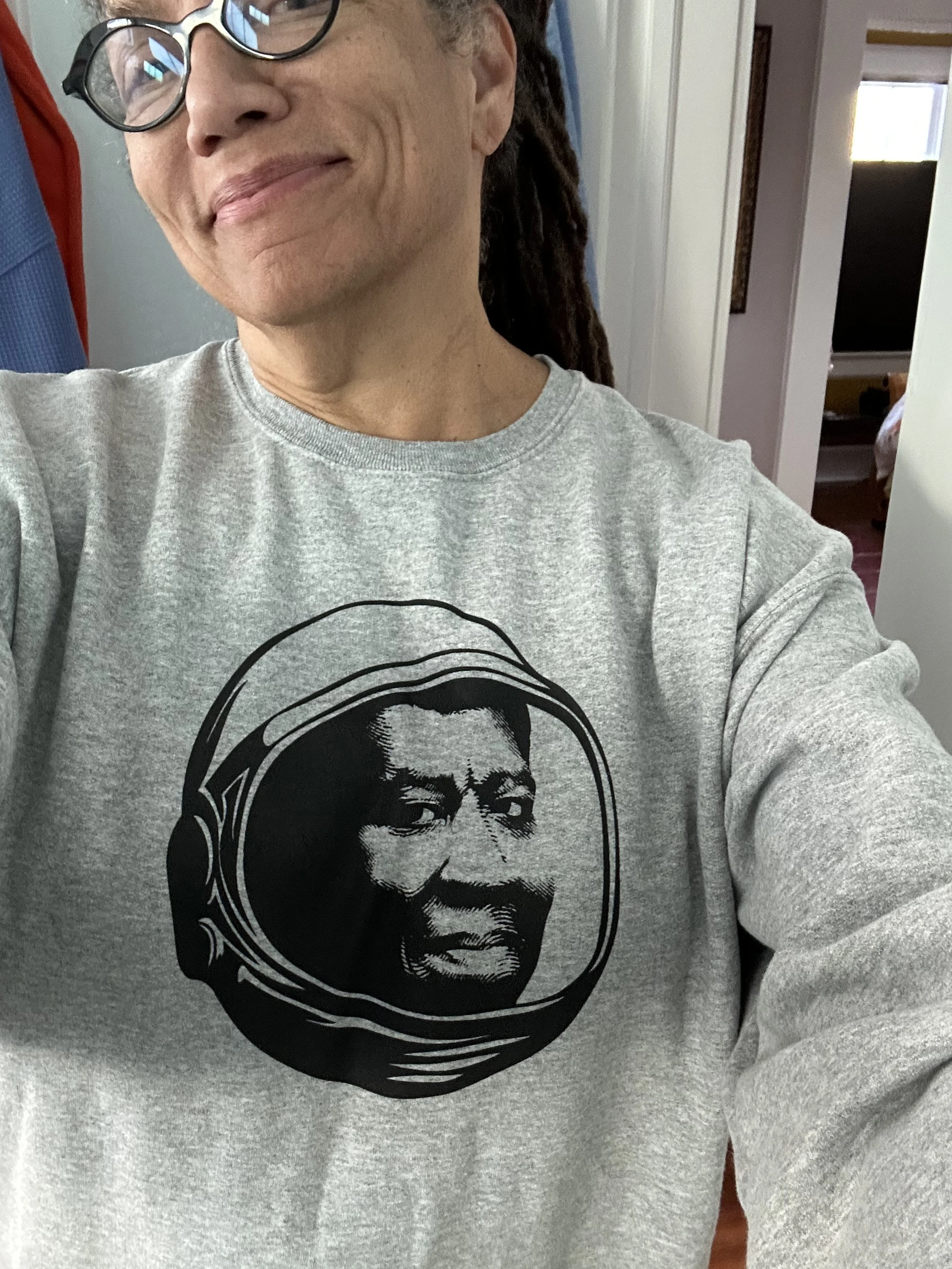 Nikky Finney with glasses smiling, wearing a gray sweatshirt with a black and white graphic of a person's face on it, indoors near a doorway.