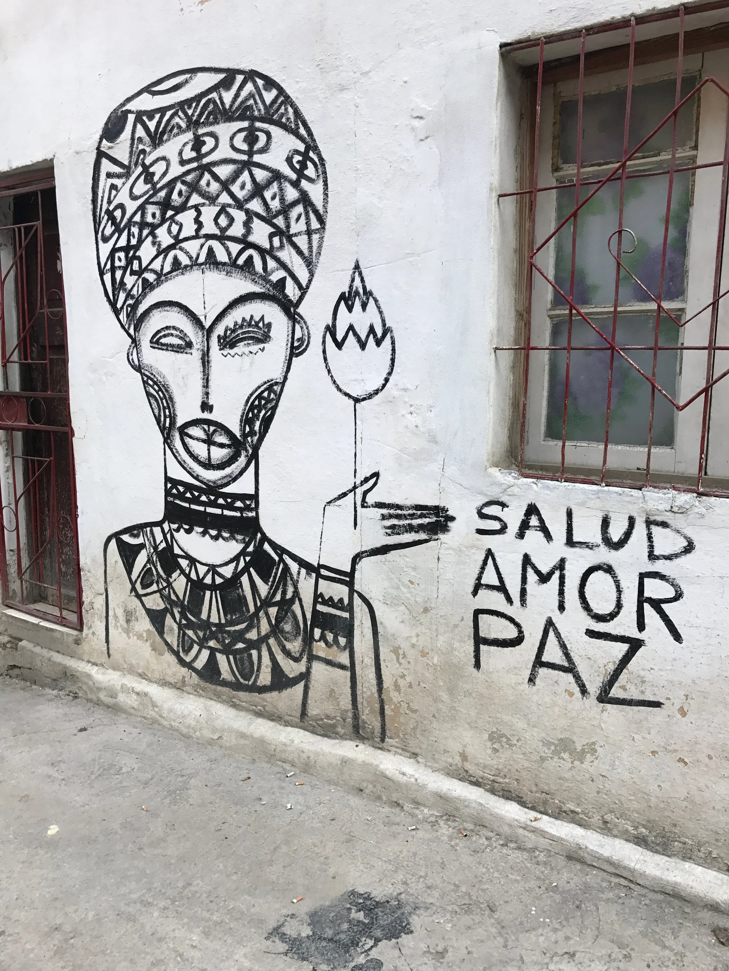Black line drawing of a smiling woman with traditional African adornments, holding a flower, written in black paint on a white wall beside a window with a red metal grid. The writing reads 'Salud, Amor, Paz'.