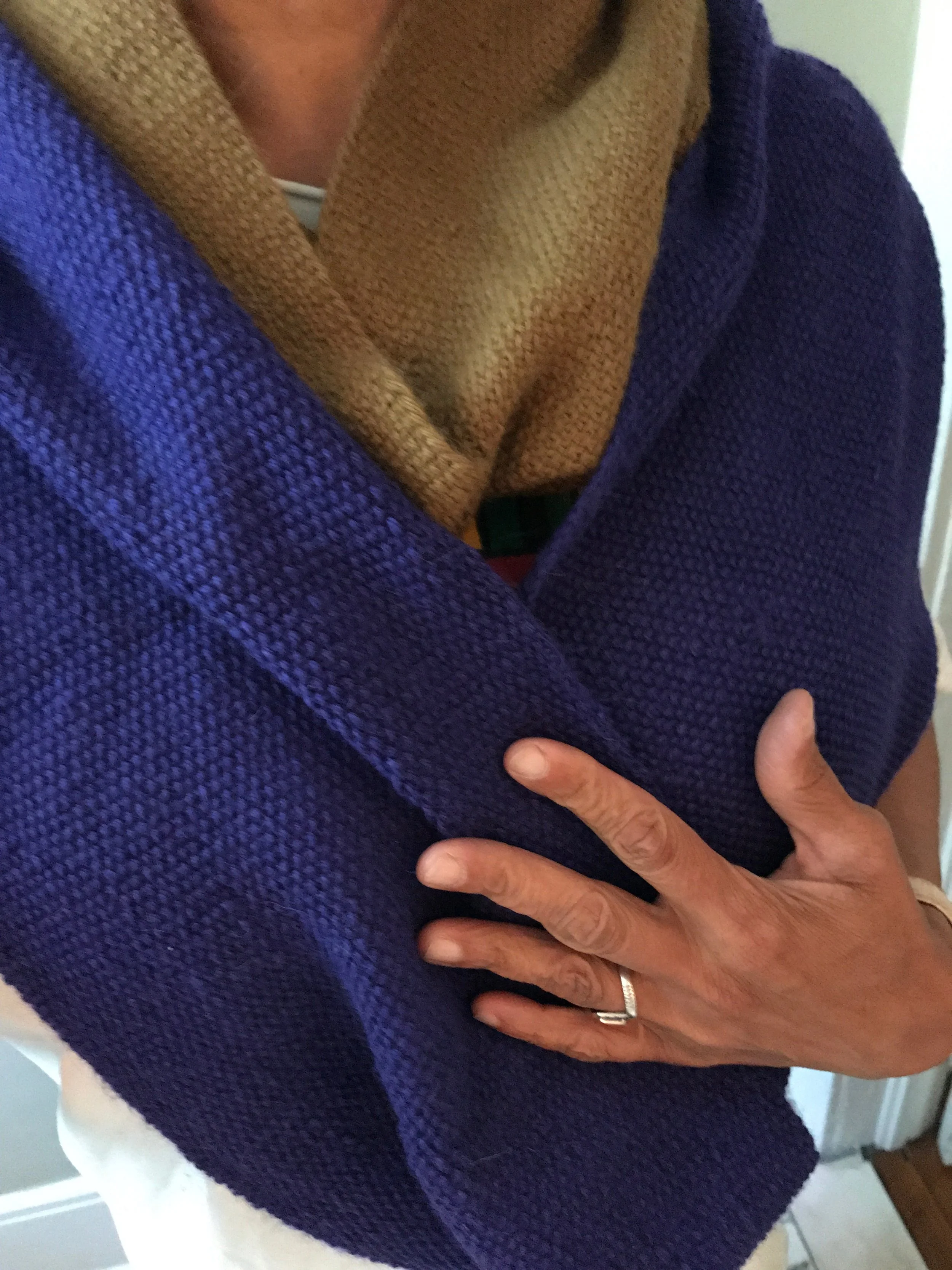 Close-up of a person wearing a purple textured cardigan and a beige scarf, with their hand resting on their chest, showing a wedding band.