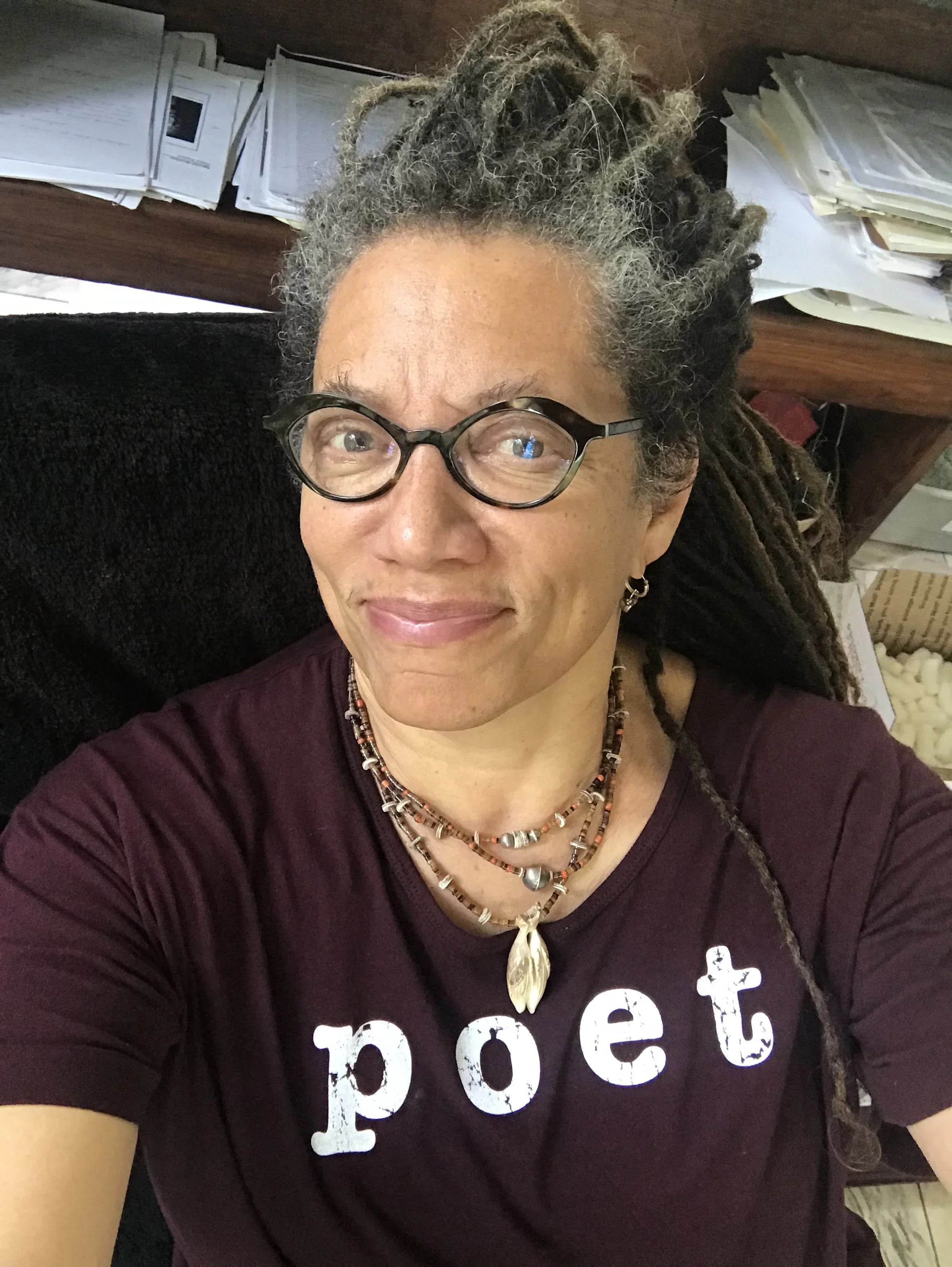 Nikky Finney with gray and black dreadlocks wearing glasses, a maroon shirt with partially visible white text that reads POET, and layered necklaces.