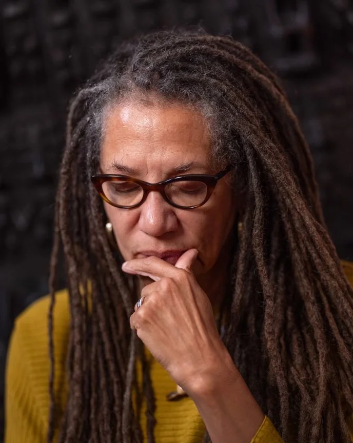Nikky Finney with long, gray dreadlocks and glasses, wearing a yellow sweater, looks down thoughtfully with her hand on her chin.