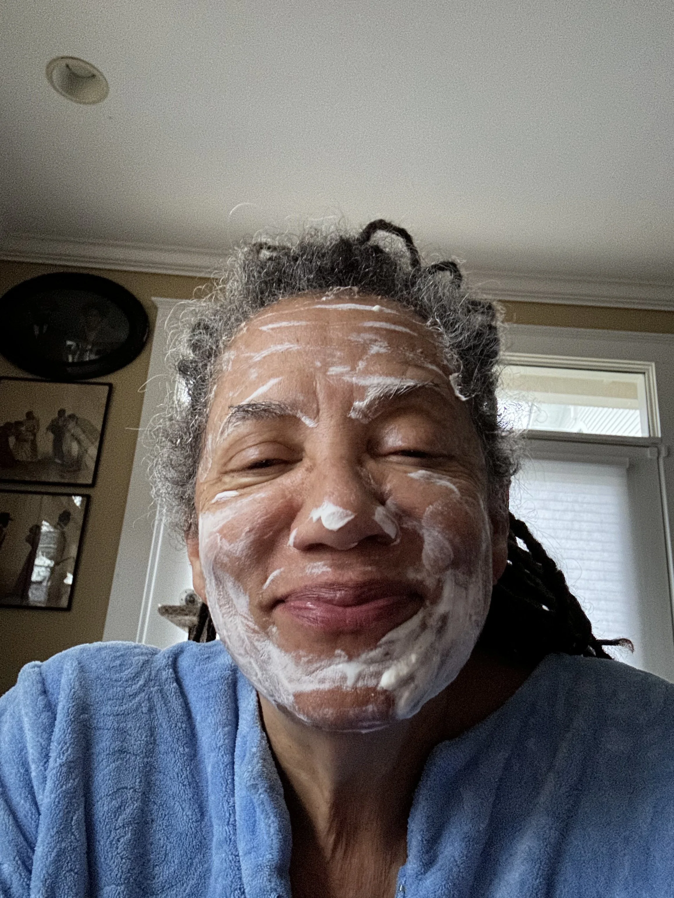 Nikky Finney with salt-and-pepper hair wearing a blue robe and smiling while washing their face with soap, indoors with windows and framed pictures on the wall.