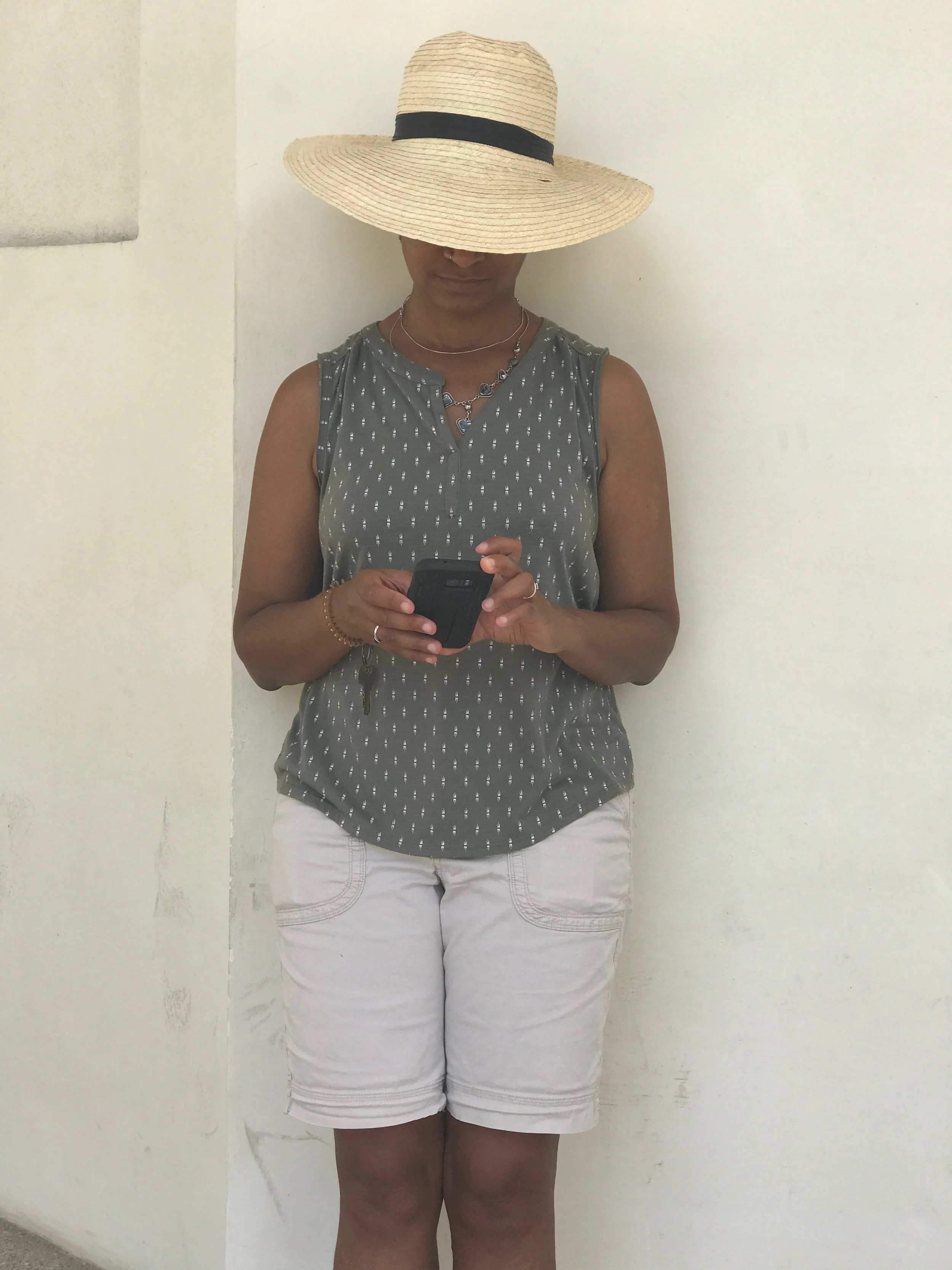 Person wearing a large straw sun hat with a black band, a sleeveless patterned top, white shorts, and holding a smartphone.