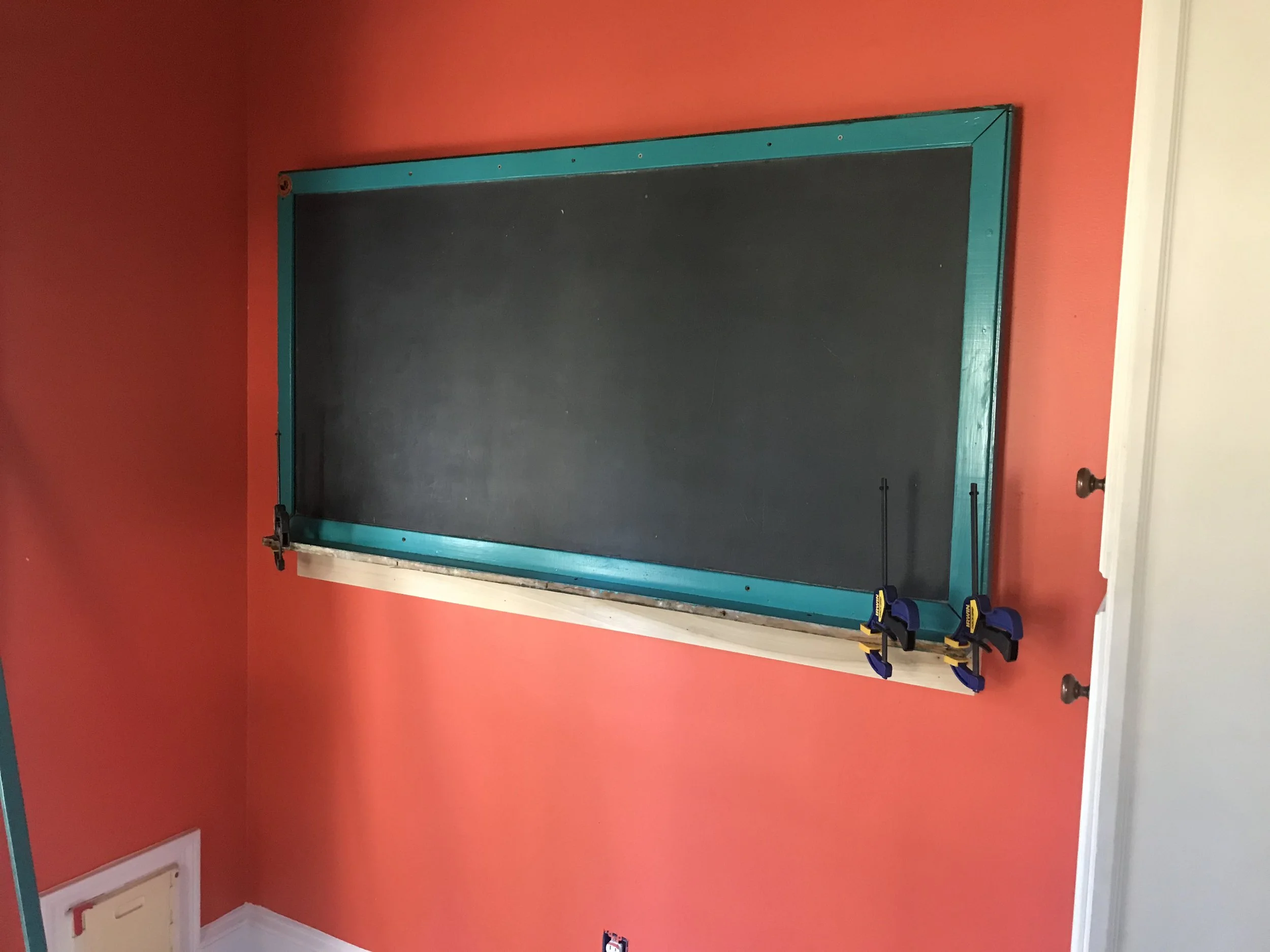 A room with red walls and white trim, featuring a blackboard with a teal-colored frame and some Clamps resting at the bottom right corner.