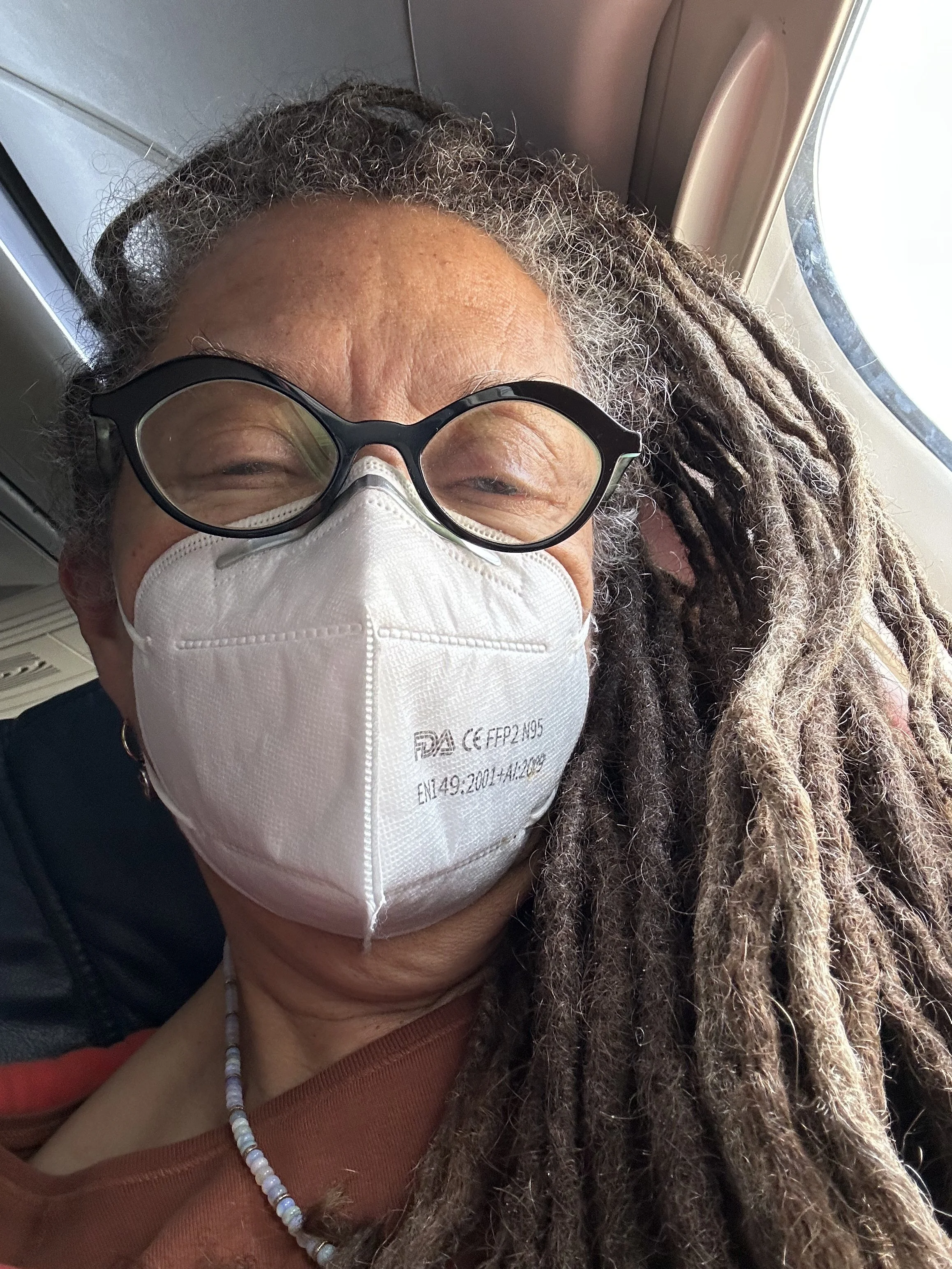 Close-up of a person with dreadlocks and glasses wearing a face mask, sitting in an airplane window seat.