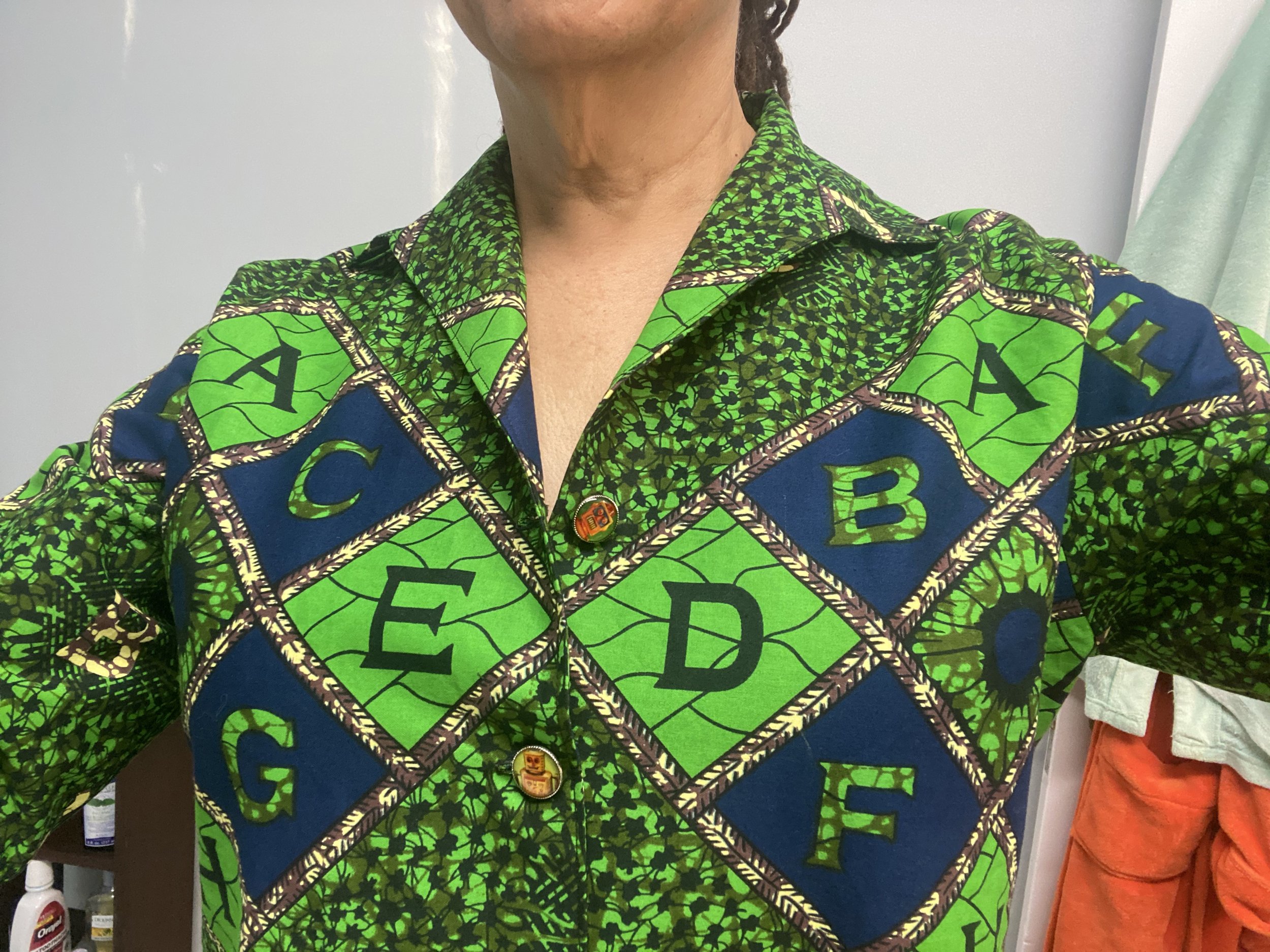 Nikky Finney wearing a colorful patterned shirt with green, blue, and black geometric designs, including the letters A, B, C, D, E, and F on the shirt. The person has light skin and dark curly hair tied back.