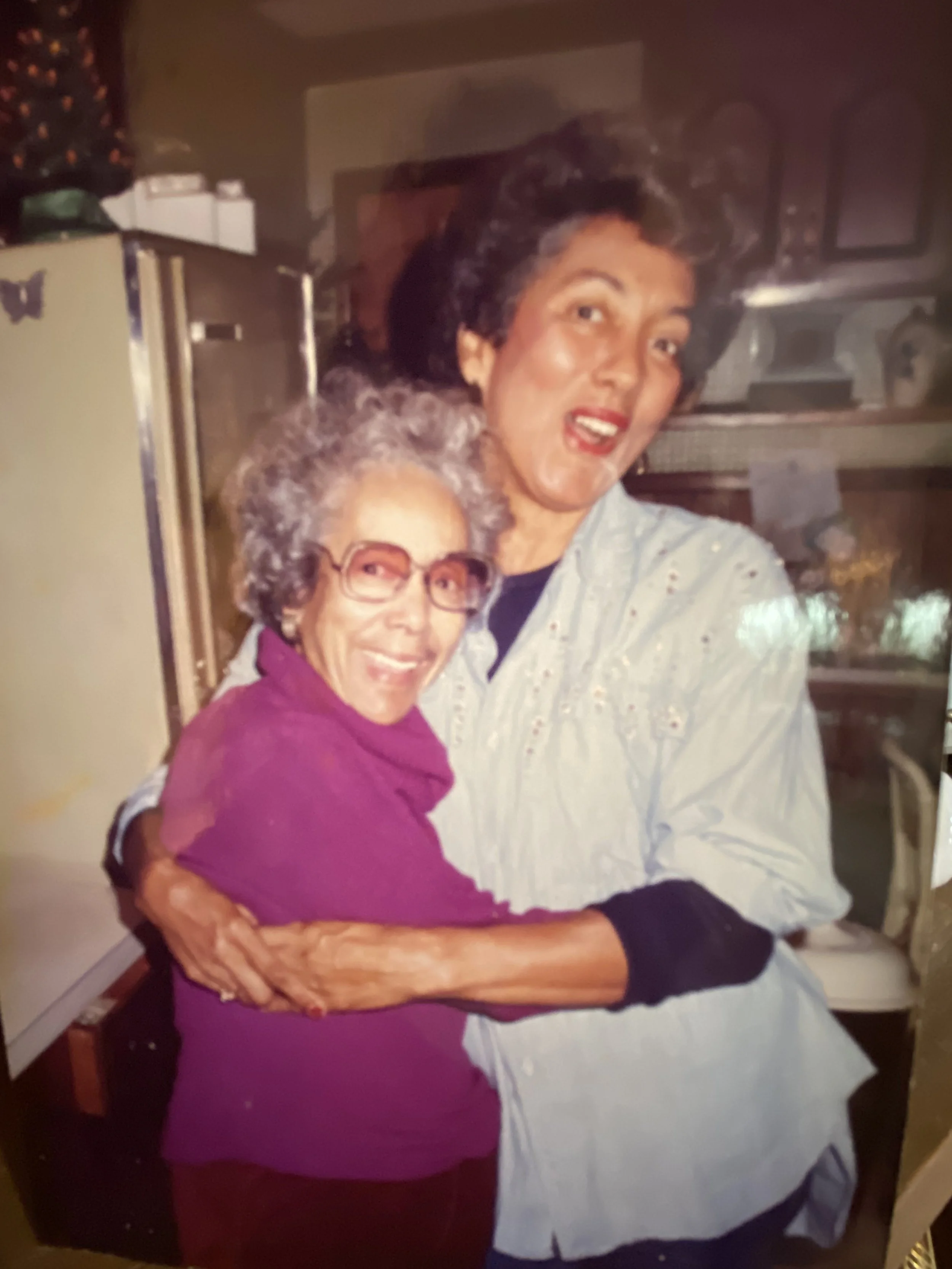 A woman hugging an elderly woman in a kitchen. The elderly woman is wearing glasses and a purple top, while the younger woman has short dark hair and is wearing a light-colored shirt with small patterns.