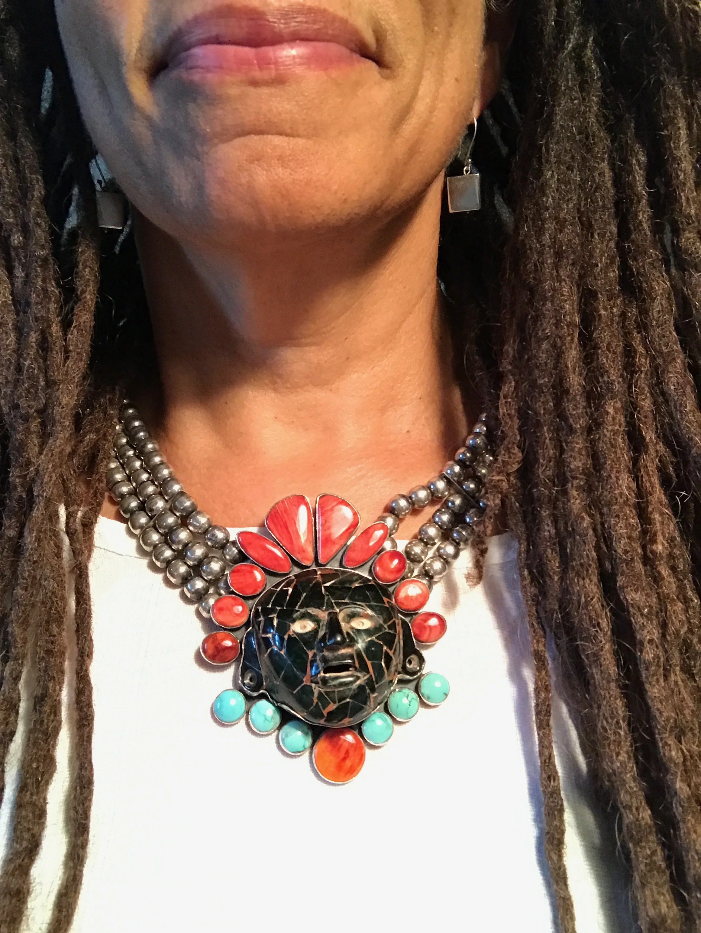 Close-up of a woman’s neck and lower face, wearing layered jewelry including a large black and orange butterfly pendant, silver, red, turquoise, and black necklaces, and an earring with a square silver charm.