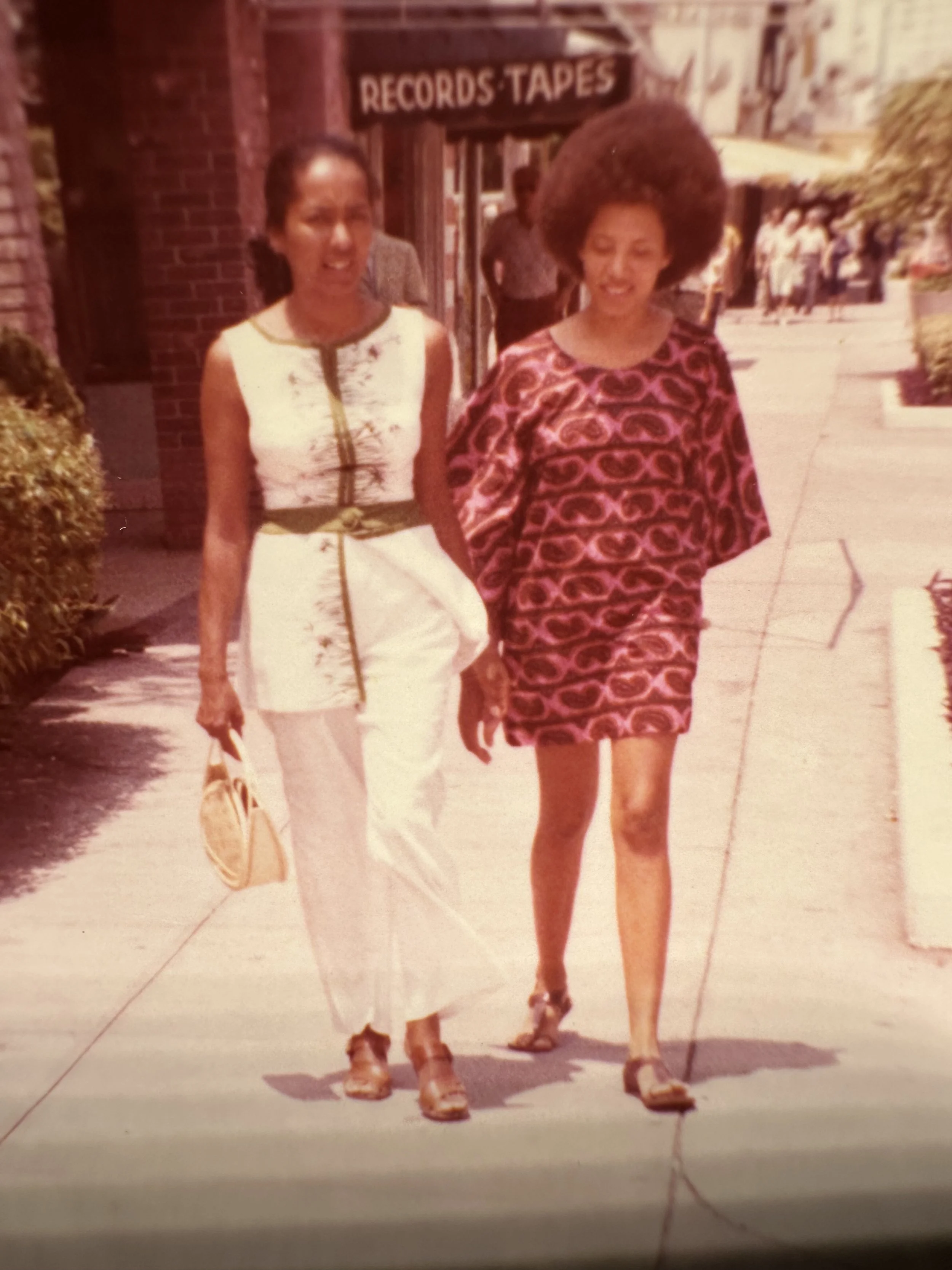 Young Nikky Finney and her mother, walking on sidewalk in front of music store with sign that reads 'Records Tapes'.