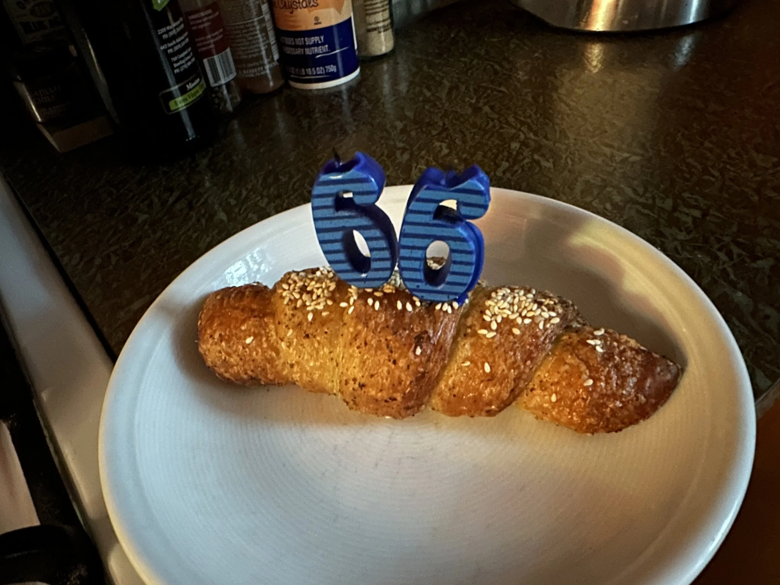 A white plate with two pastries topped with sesame seeds and blue number candles reading '66'. Background includes spice bottles and a stainless steel container in a kitchen counter.