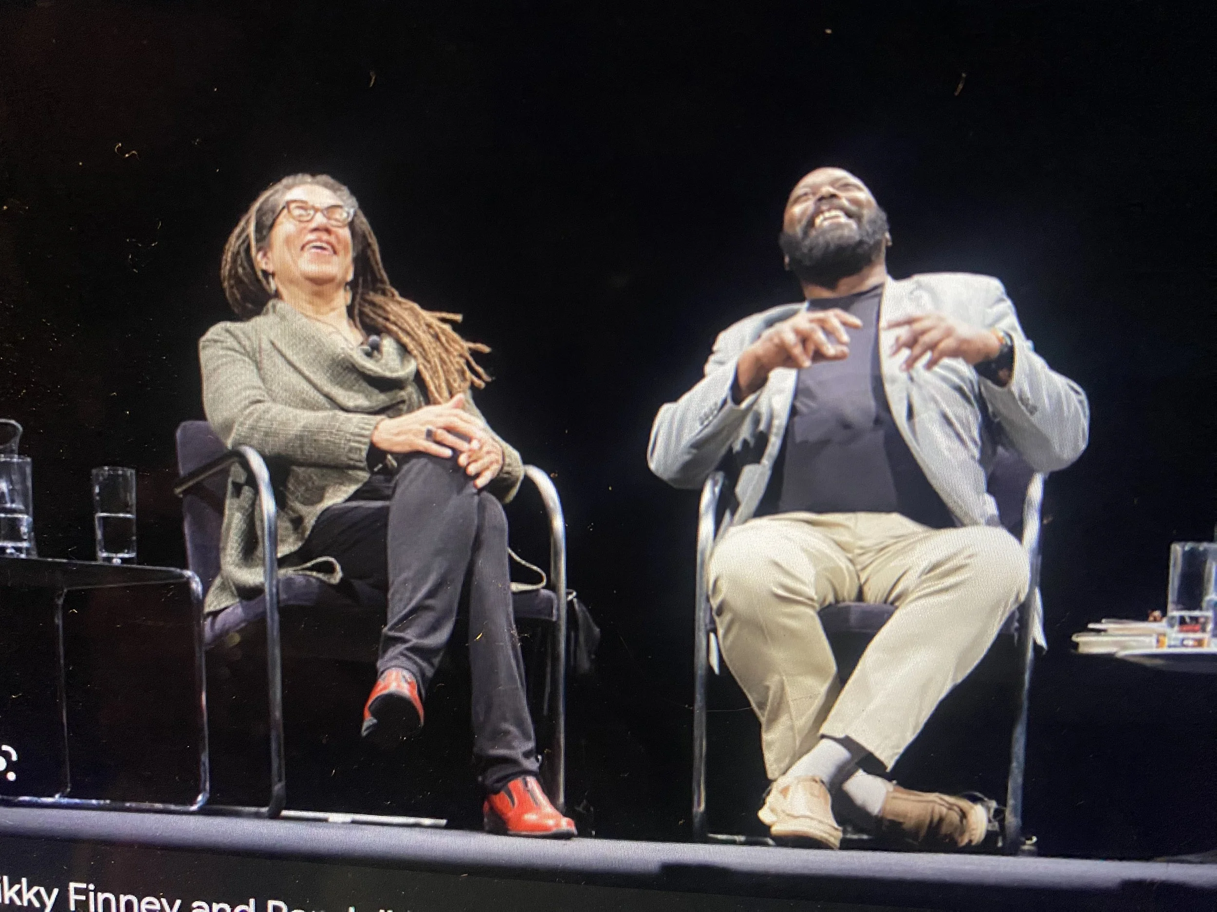 Two people are sitting on chairs against a black background. The woman on the left has dreadlocks, glasses, and is wearing a beige jacket. The man on the right has a beard and is wearing a light-colored blazer and beige pants. Both are smiling and la