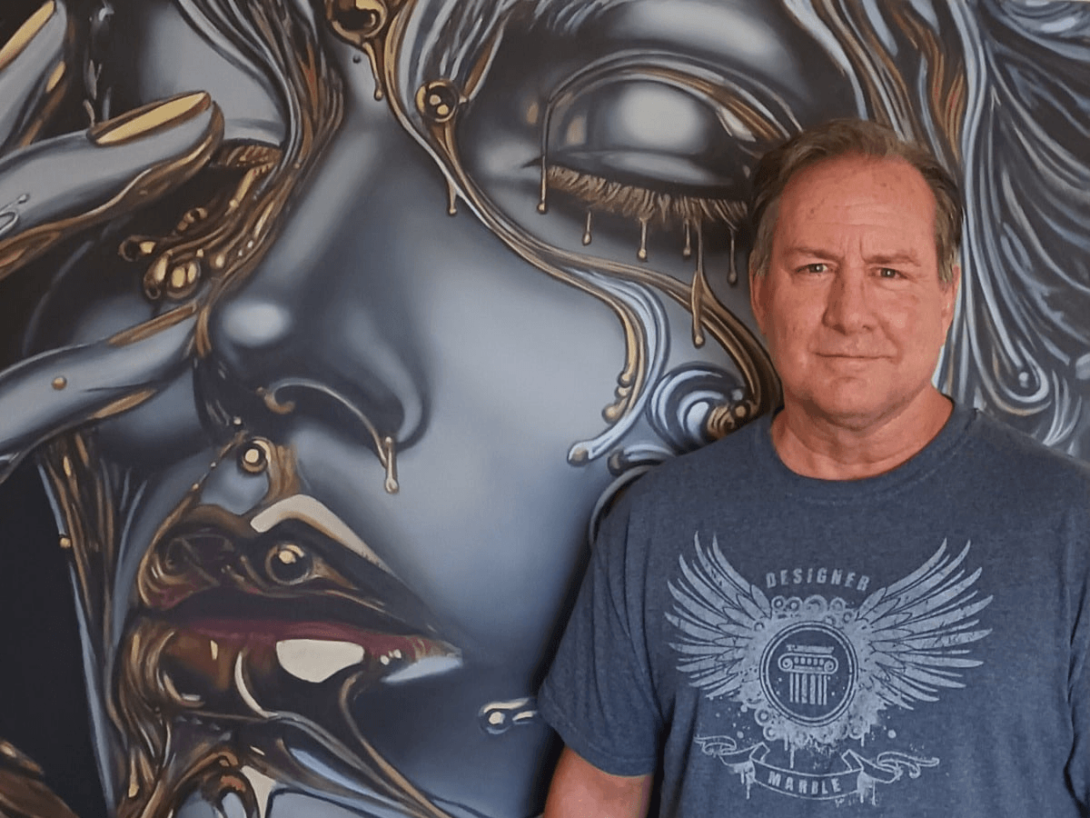 A man standing in front of a large artistic mural of a woman's face painted in metallic colors with gold and silver accents.