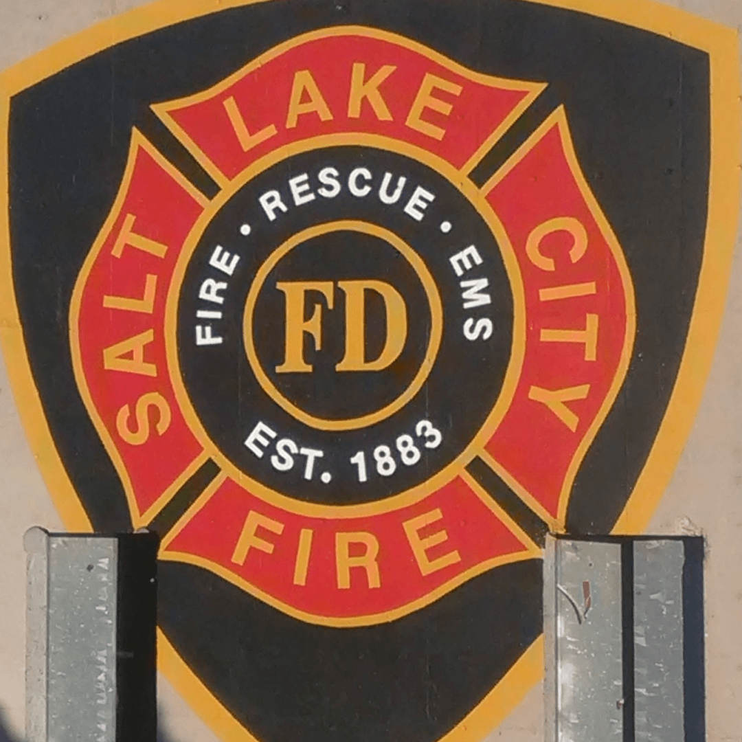 The shield-shaped emblem of the Lake City Fire Department, established in 1883, with a black background, red and yellow text and design elements, and the initials 'FD' in the center.
