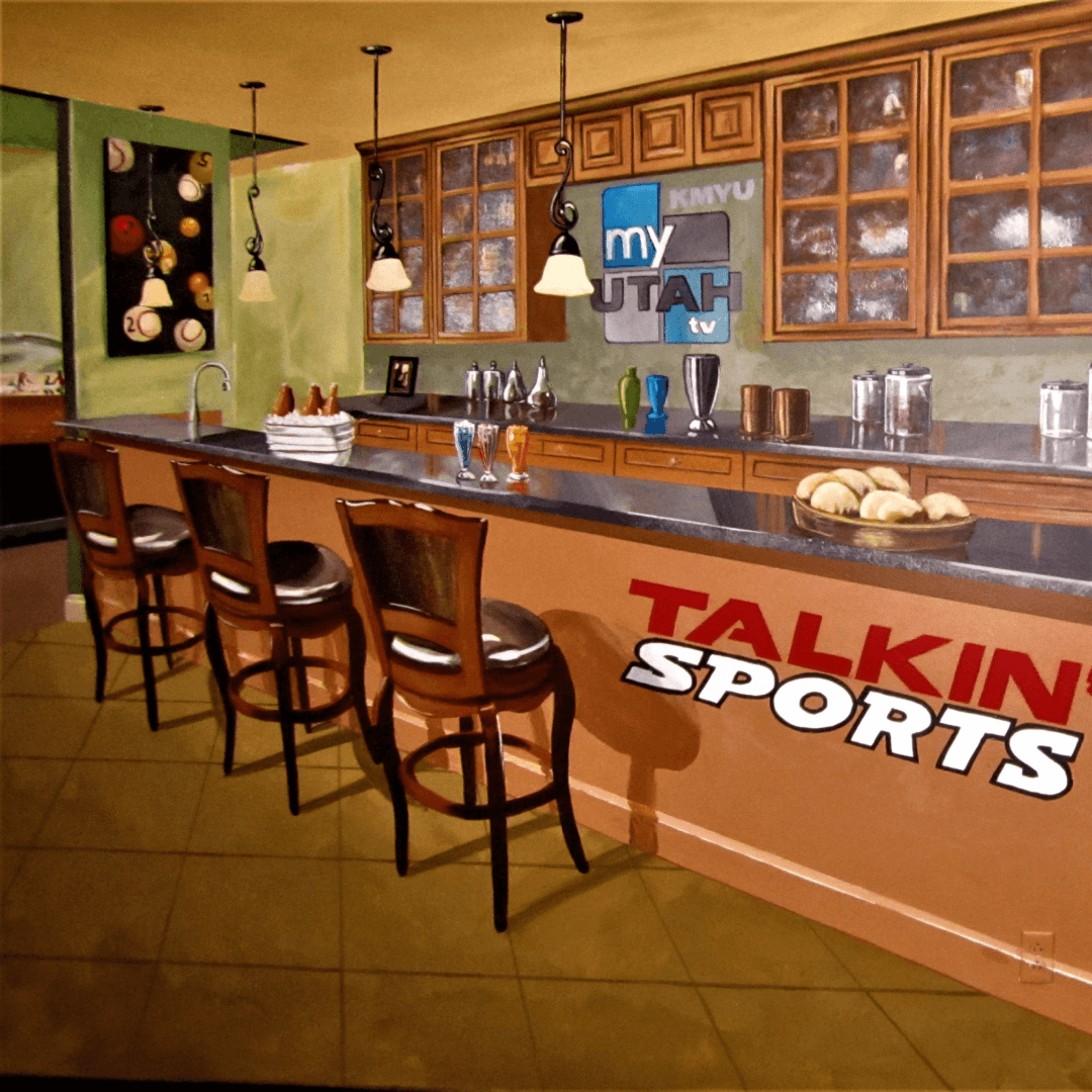 A bar area with four wooden chairs, a black countertop, and glassware. There are kitchen cabinets with glass doors, various objects on the counter, and sports-themed décor, including tennis balls and a framed sports display. A sign on the wall reads 