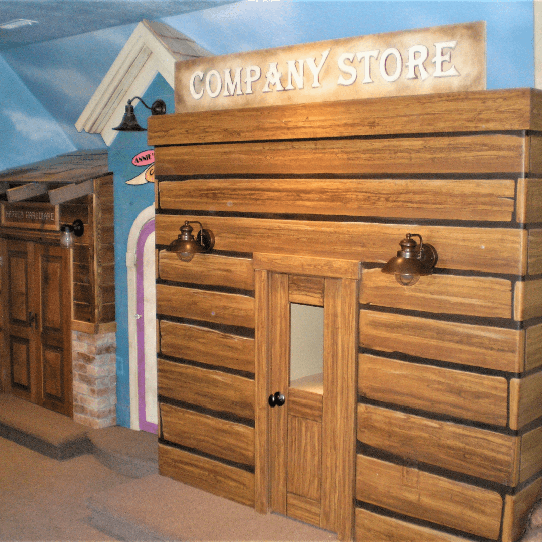 A miniature store resembling a rustic company store with a wooden facade, two wall-mounted lamps, and a sign reading 'Company Store' above the entrance.