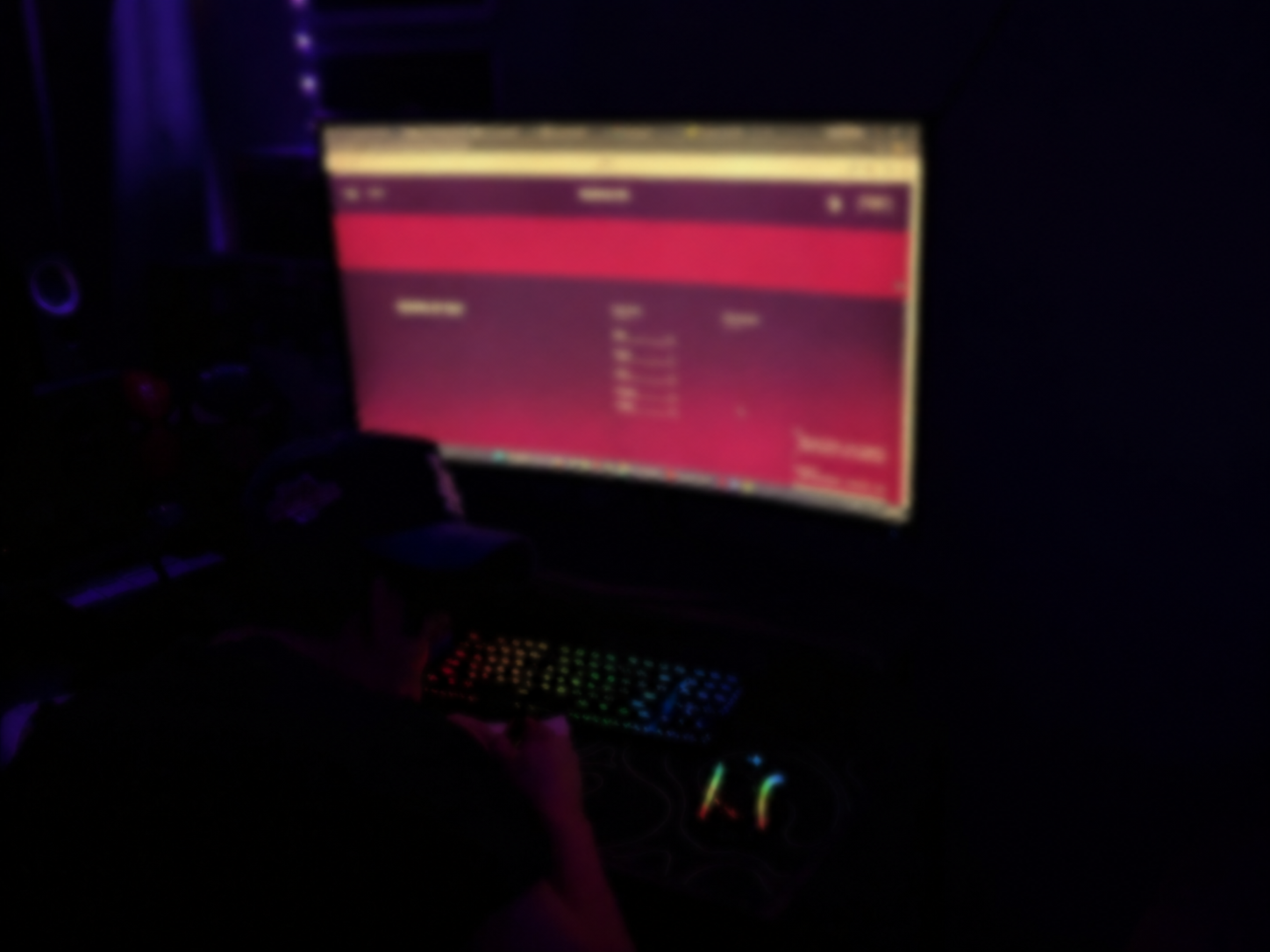 A dimly lit room with a computer monitor displaying a red and purple screen, a person using a keyboard with rainbow-colored backlighting, and various equipment on a desk. Web design.