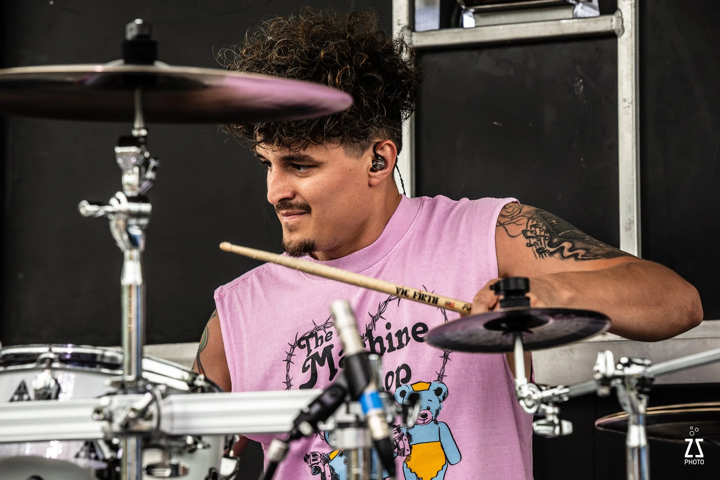A young man with curly hair playing a drum set in a music rehearsal space.