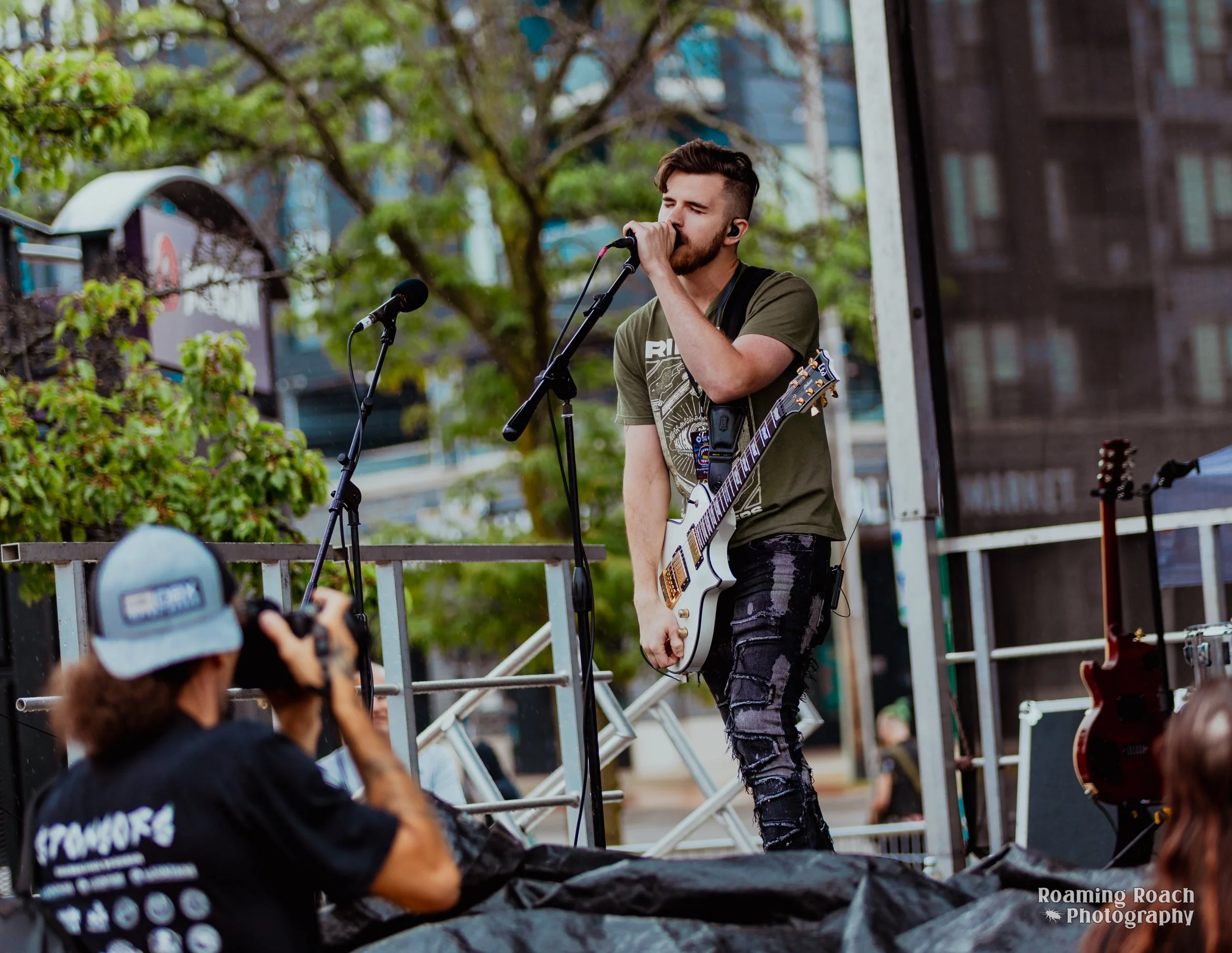 A young male musician with styled hair and a beard performs outdoors on a small stage, singing into a microphone while holding an electric guitar. The background shows trees and a cityscape. Audience members are visible in the foreground, with one person taking a photo.