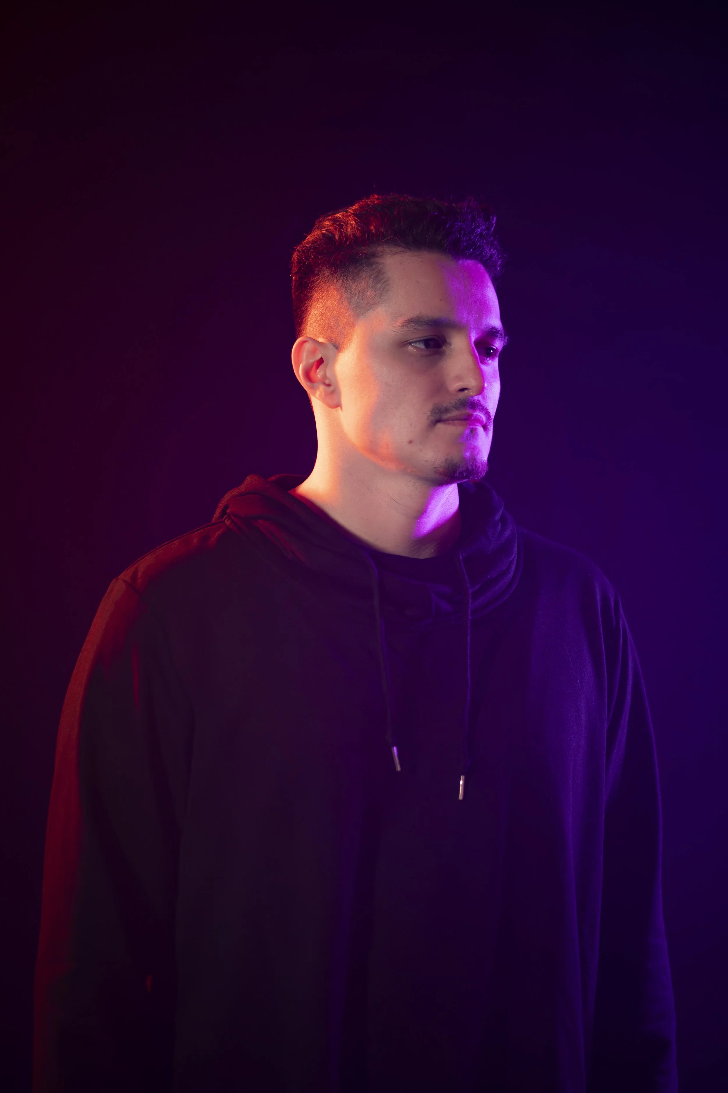 A young man with short dark hair and a small goatee, wearing a black hoodie, is illuminated by pink and purple lighting against a dark background.