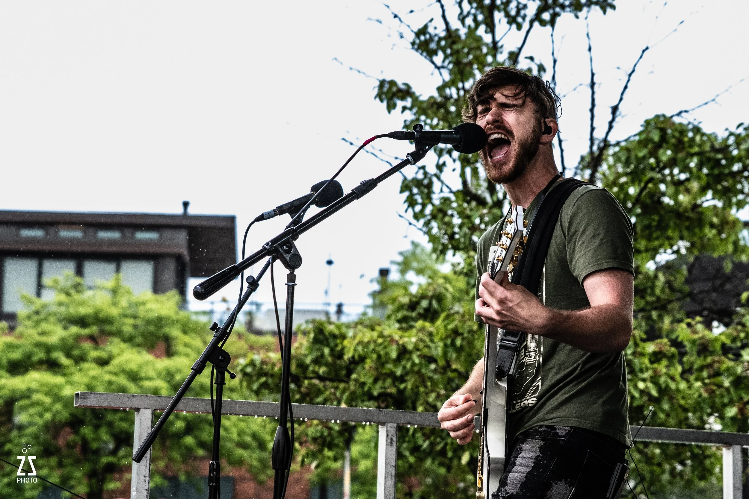A man with dark hair, beard, and wearing a green t-shirt is singing passionately into a microphone while playing an electric guitar outdoors on a cloudy day. There are trees and a building in the background.