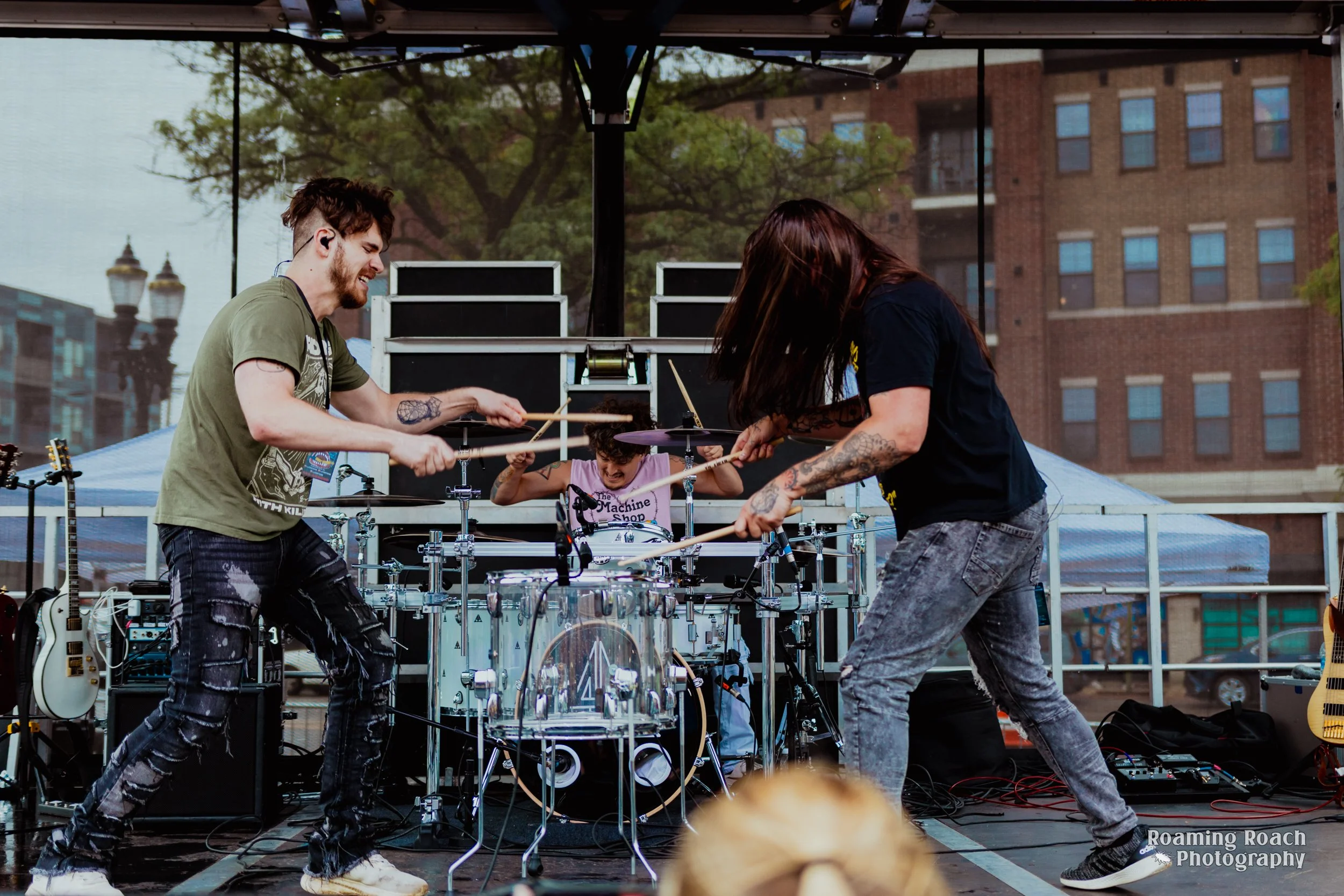 Two musicians with tattoos and casual clothing perform on an outdoor stage, playing drums and guitars, with a person in the background behind a drum set. Buildings and trees are visible in the background.
