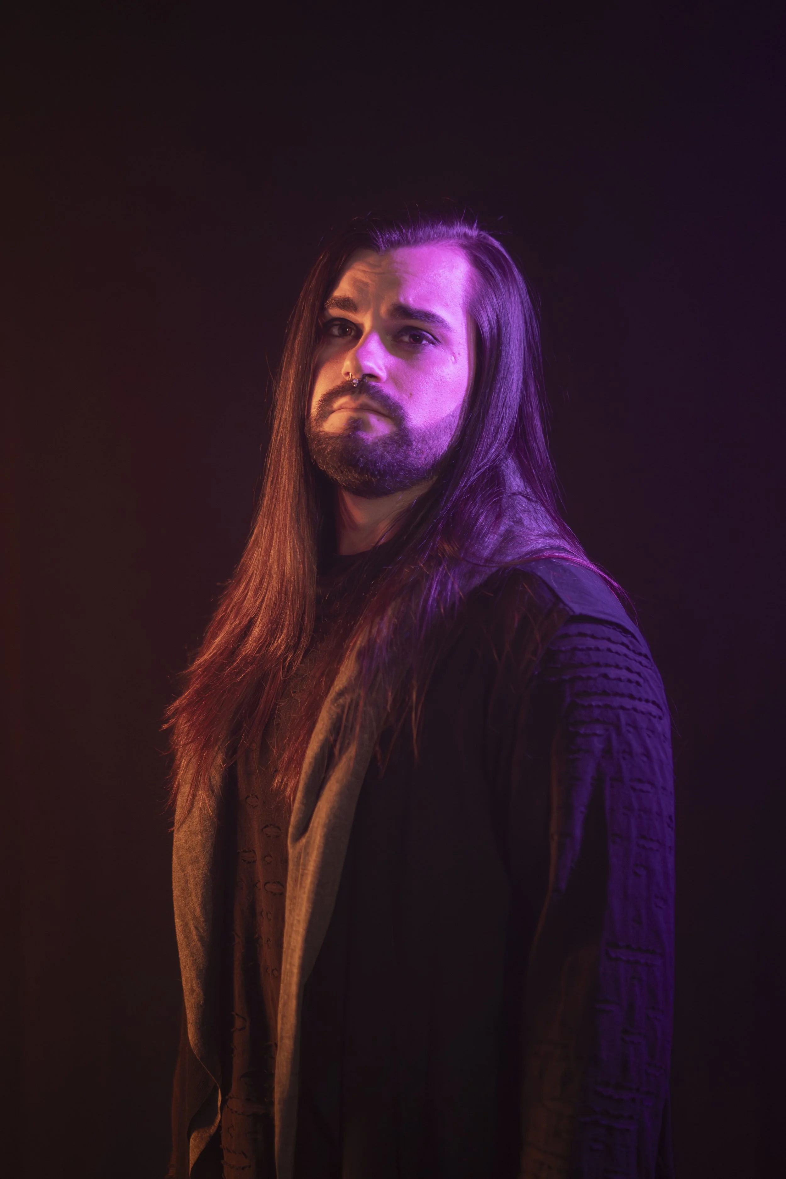Portrait of a man with long hair and a beard, illuminated by pink and purple lighting, against a dark background.
