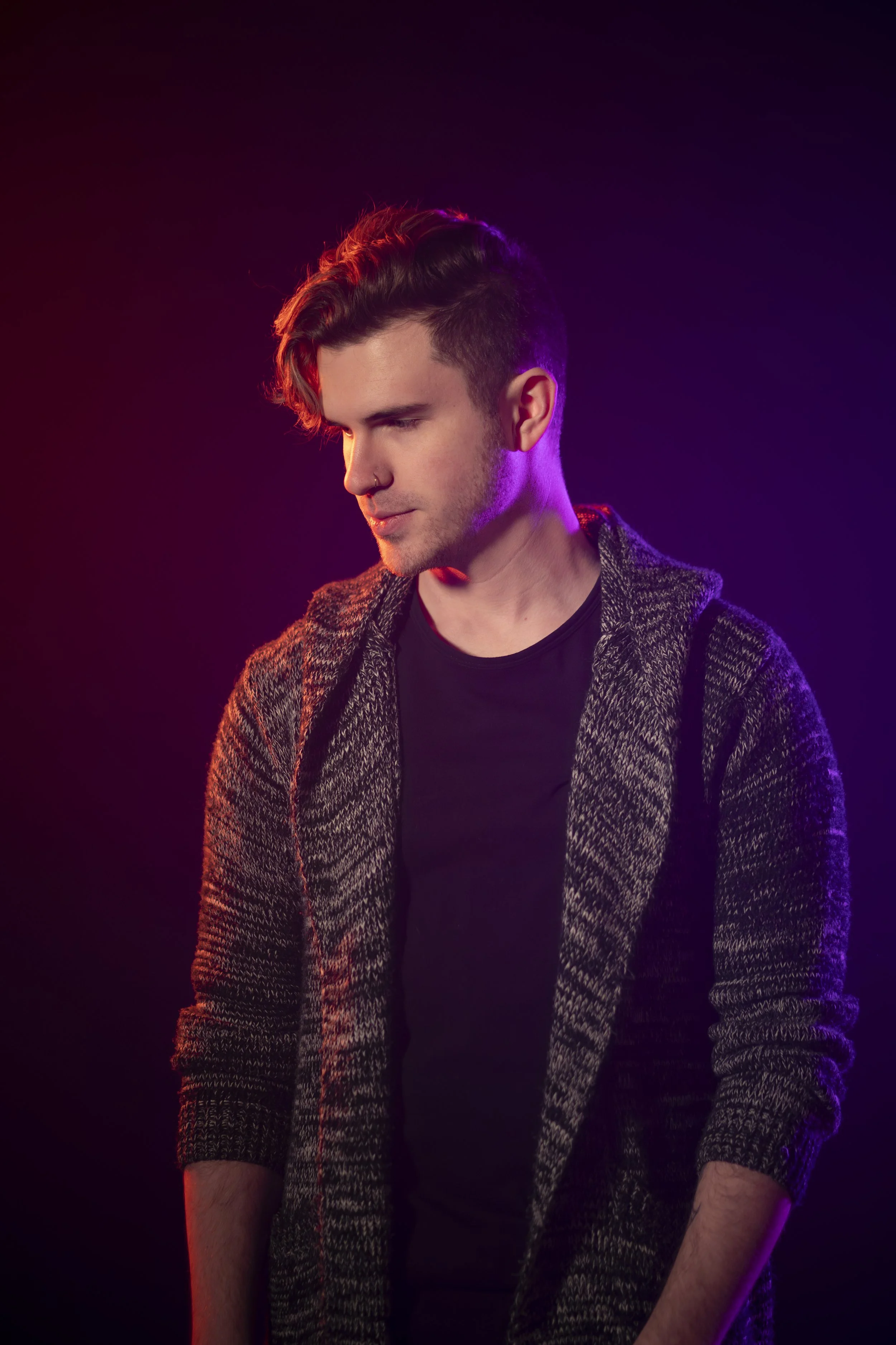 Young man with dark hair and a nose piercing, wearing a black shirt and a patterned blazer, posed against a dark background with colorful stage lighting.