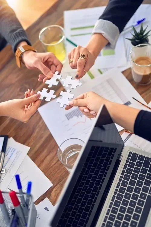 Multiple hands holding white puzzle pieces over a cluttered desk with papers, a laptop, pens, and drinks.