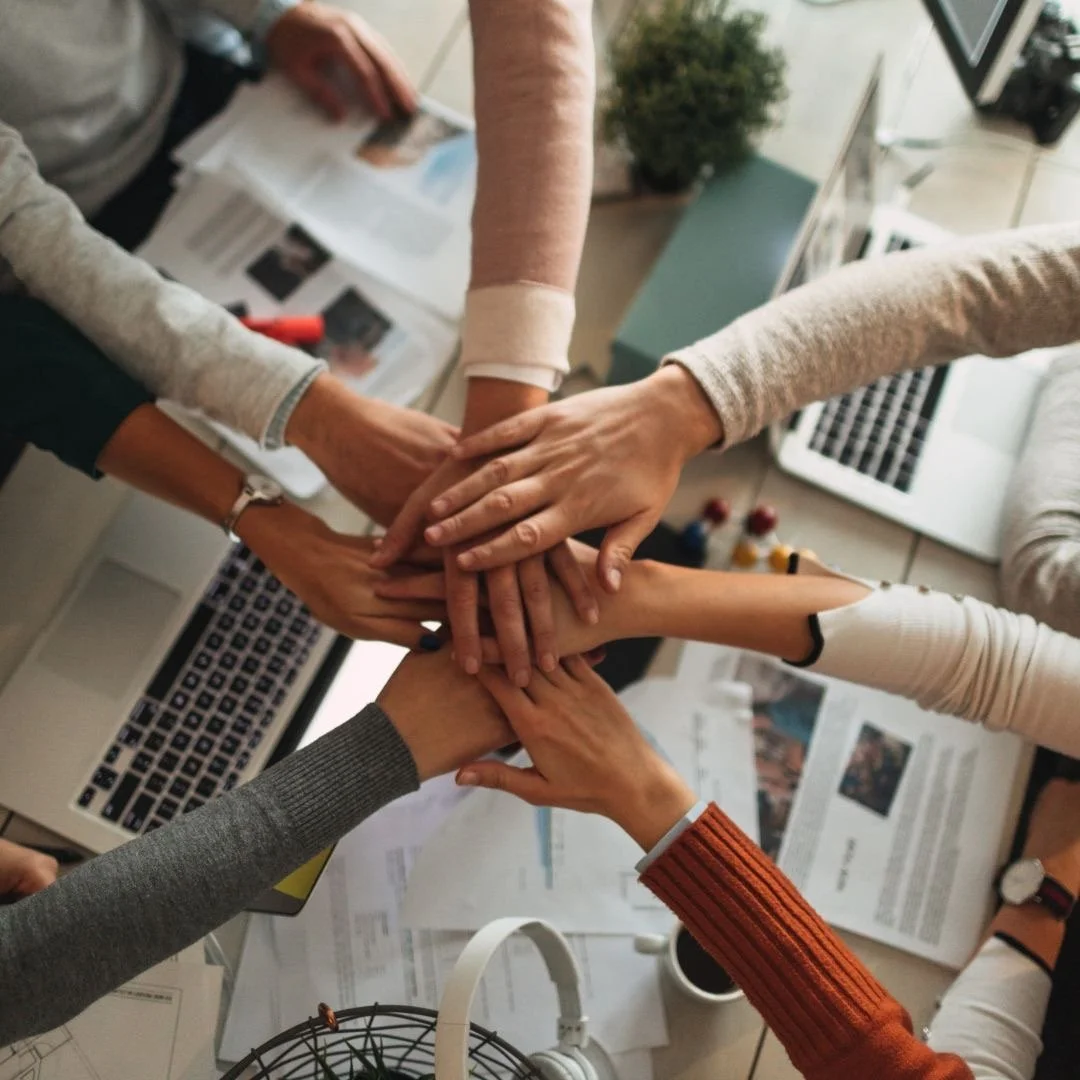 A group of diverse people place their hands together in a collaborative gesture over a cluttered desk with laptops, papers, and office supplies.
