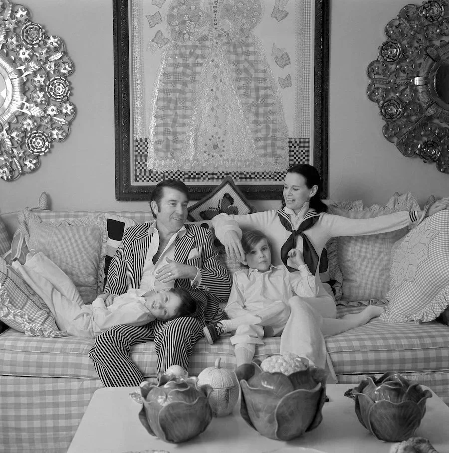 A family of four sitting on a patterned sofa in a living room, smiling and relaxing. The man is wearing a striped suit, the woman has her arms outstretched, and two children are seated between them, one lying down and one sitting upright. The room decor includes framed artwork, decorative plates on the wall, and a table with ceramic bowls and pumpkins in front.