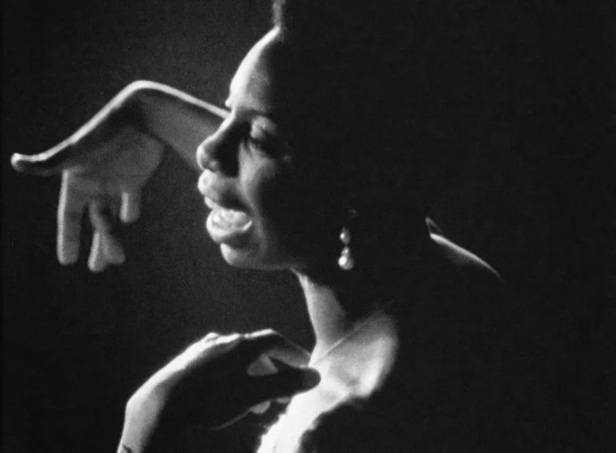 A black and white photograph of a woman singing or speaking passionately, with her eyes closed and hand near her face, wearing a earring and an elegant necklace.
