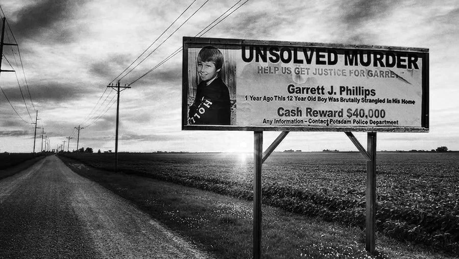 A large billboard on the side of a rural road displaying an unrewarded reward for information on the murder of Garrett J. Phillips, a 12-year-old boy. The billboard features a photo of Garrett and details about his brutal death and the reward amount.