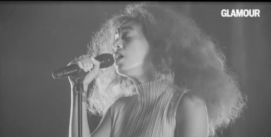 Black and white photo of a woman singing into a microphone with her eyes closed, with the word 'Glamour' in the top right corner.