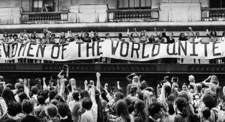 A large group of women holding a banner that reads 'WOMEN OF THE WORLD UNITE' at a protest or rally in front of a government building, with an audience of diverse people raising their fists and listening.