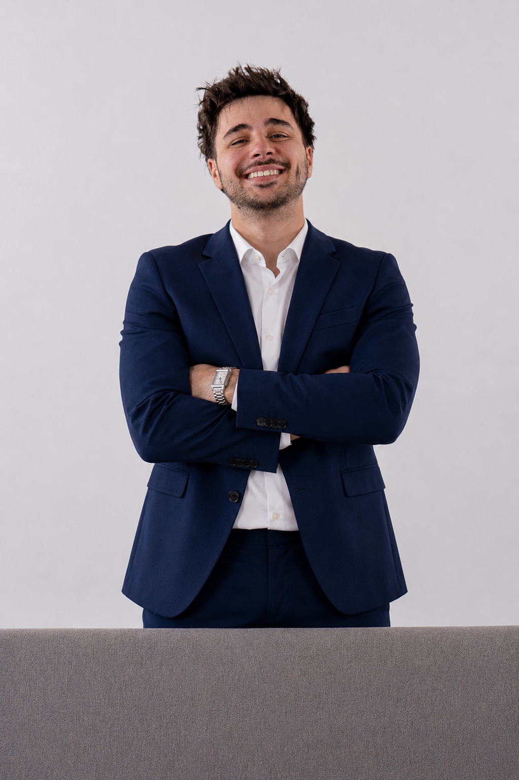 A young man with dark brown hair and beard, wearing a navy blue suit and white shirt, smiling with arms crossed, standing behind a gray table against a plain light gray background.