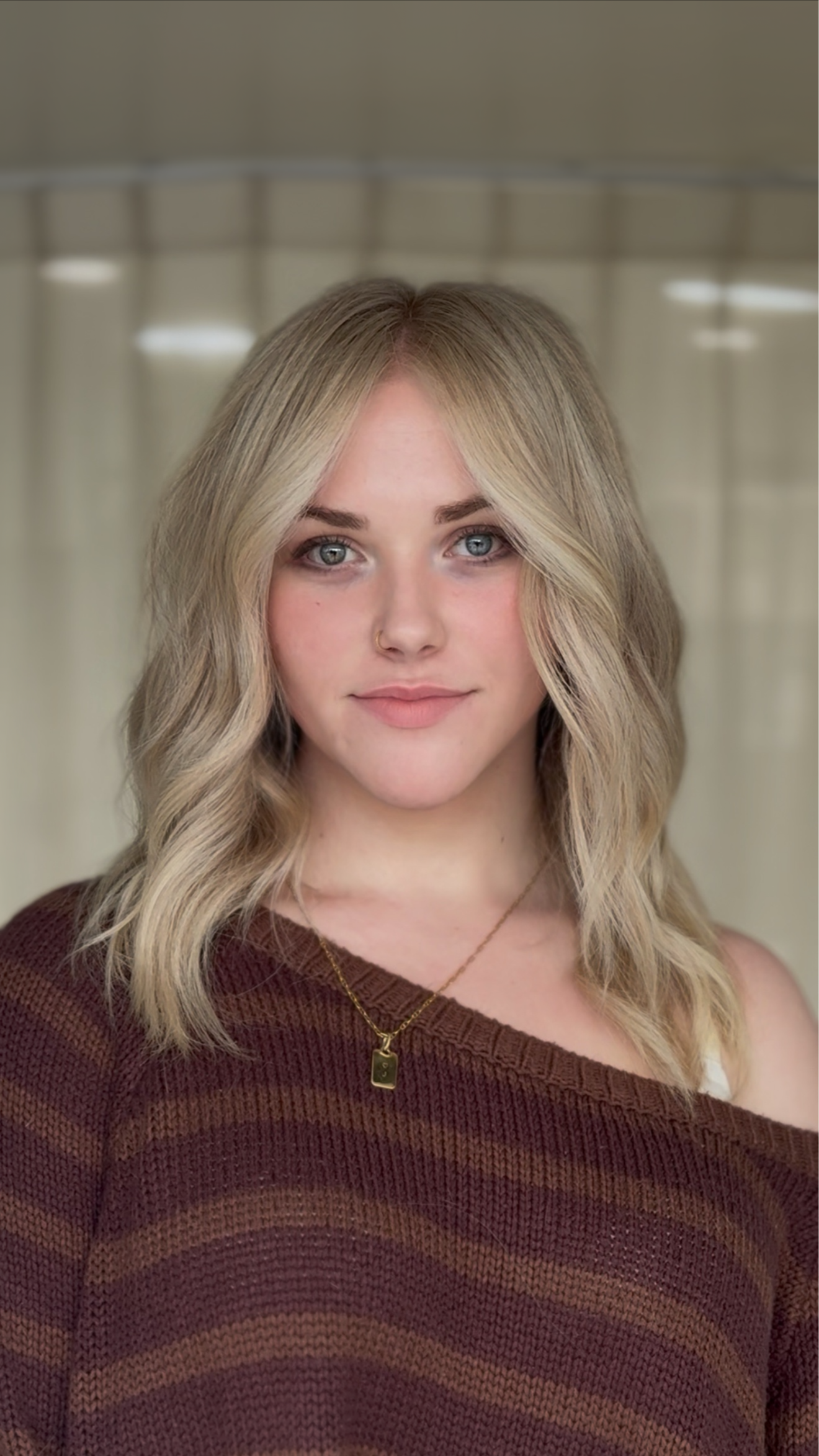 A young woman with blonde wavy hair and blue eyes wearing a maroon and tan striped sweater and a gold necklace with a pendant.